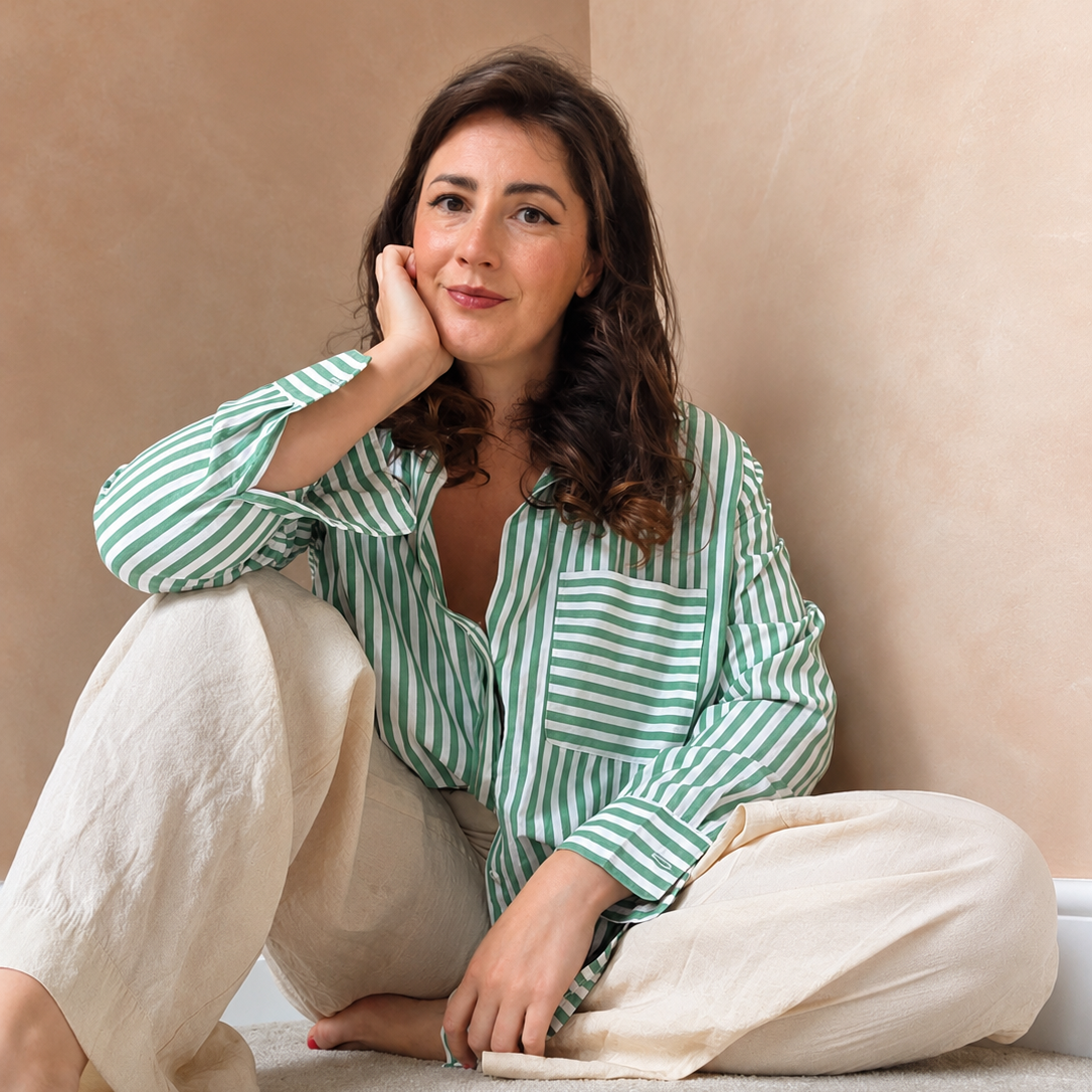 Woman with wavy brown hair wearing green and white striped shirt and beige pants, sitting on the floor and resting her head on her hand.