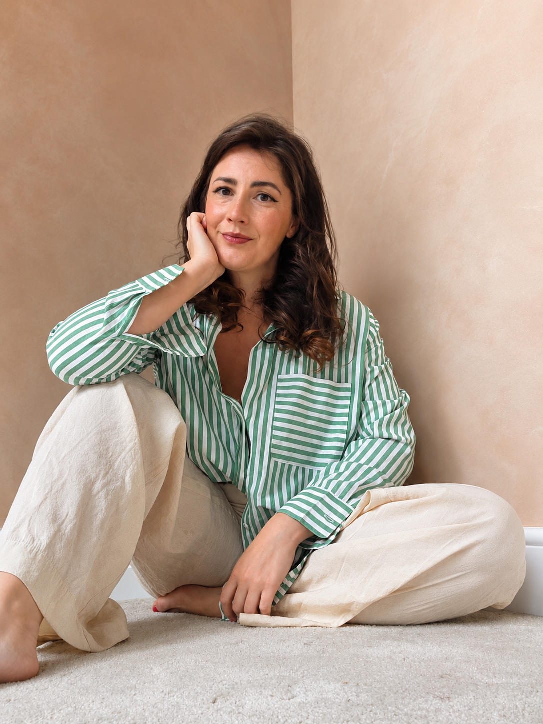 A woman with brown, curly hair wearing a green and white striped shirt and beige pants, sitting on a light-colored carpeted floor, resting her head on her hand, in a softly lit room with beige walls.