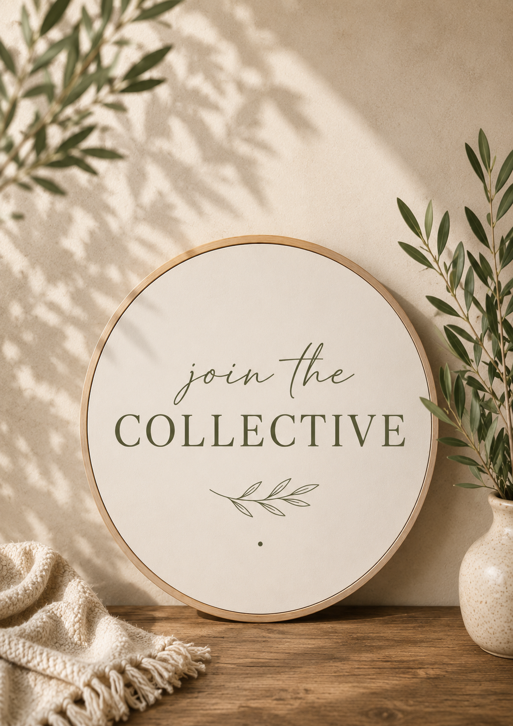 Round sign with the text 'join the COLLECTIVE' surrounded by green plants and a beige cloth on a wooden surface.