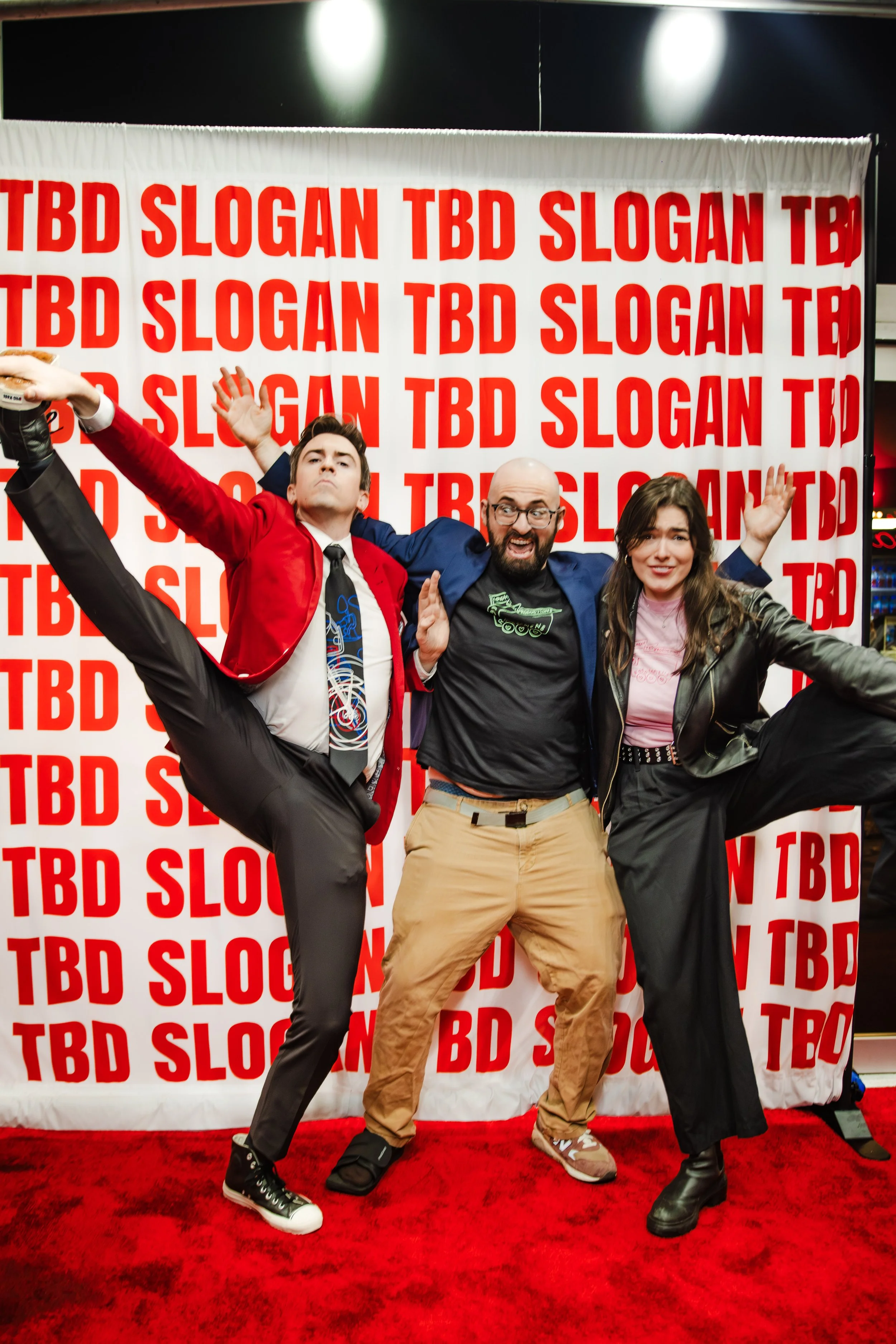 Two men and a woman strike crazy poses on a red carpet.