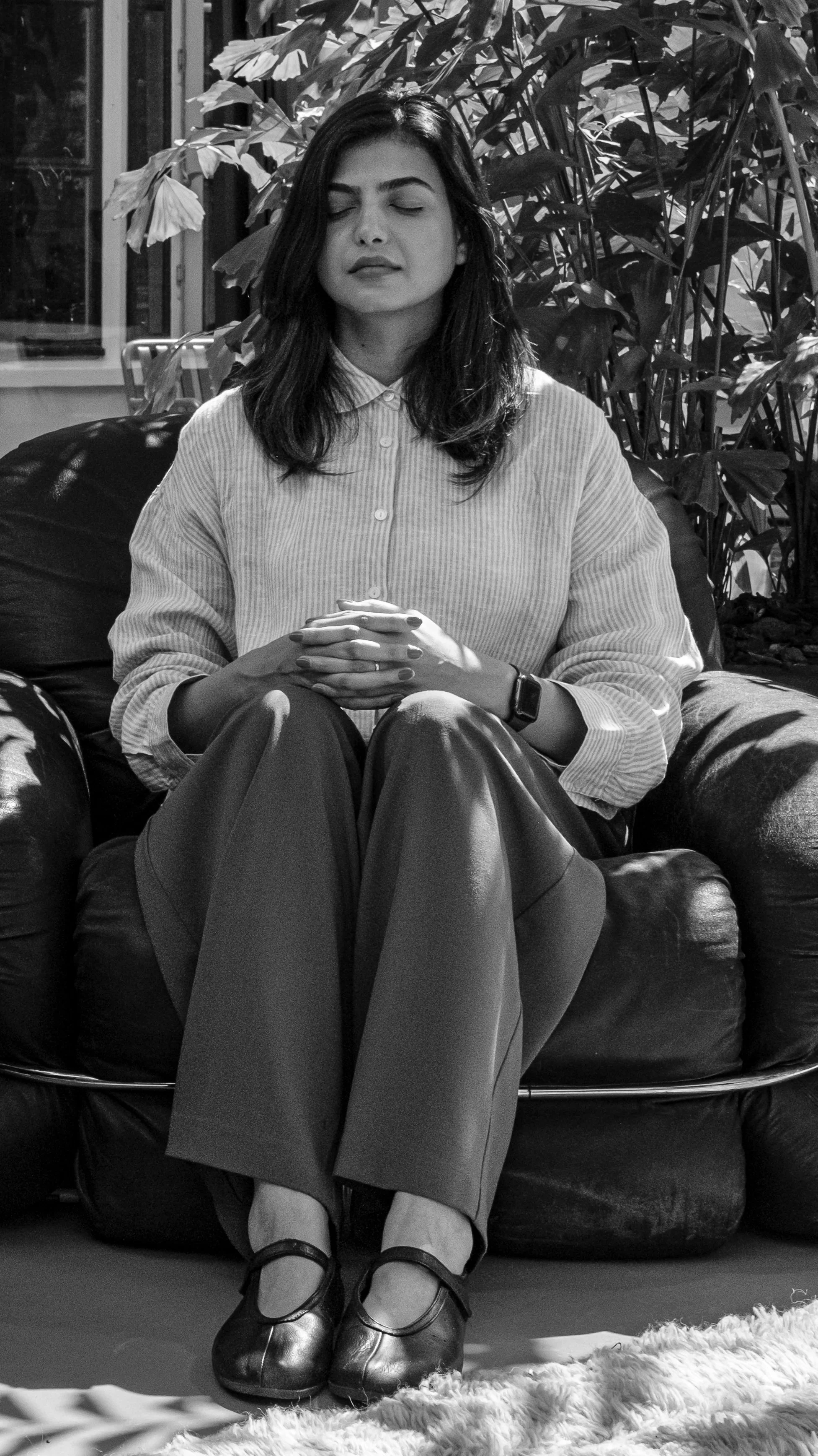 A woman with closed eyes sitting on a leather couch with her hands resting on her lap, wearing a collared shirt and trousers, in front of a large plant in an indoor setting.