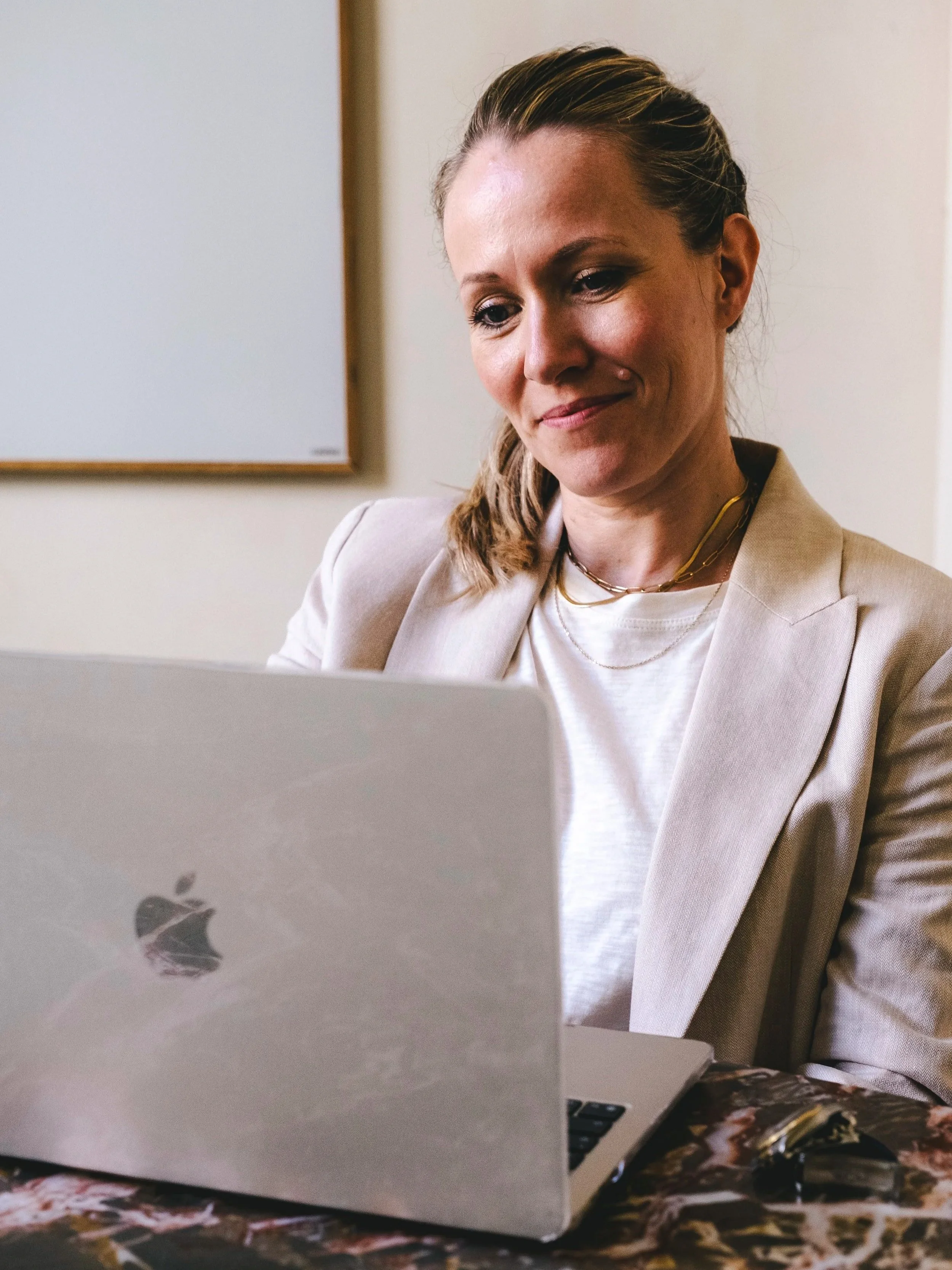 A woman with blonde hair pulled back, wearing a beige blazer and white top, sitting at a desk and looking at a silver MacBook laptop, with a whiteboard on the wall behind her.
