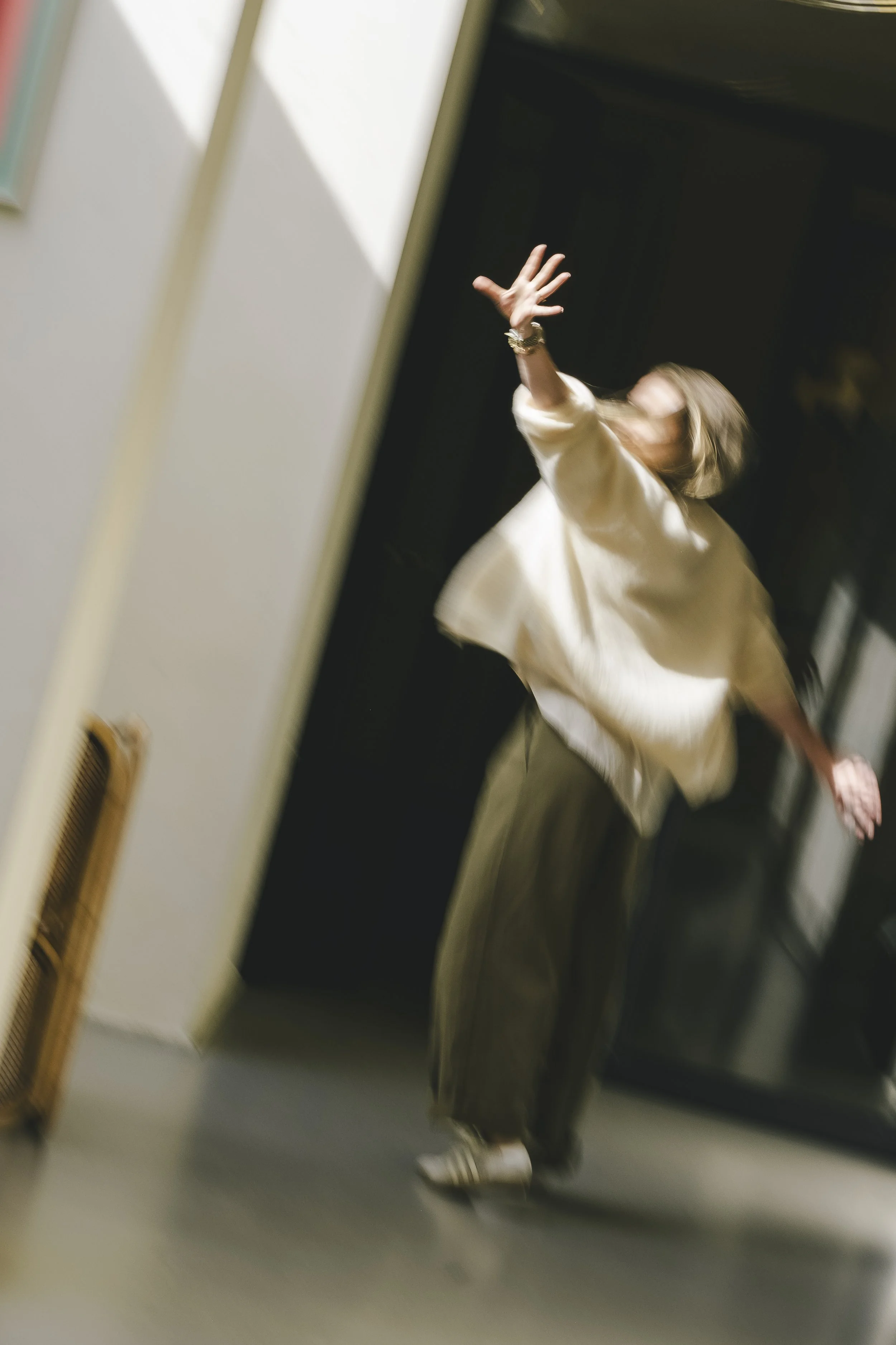 Blurred photo of a person with blonde hair reaching out with their right hand, wearing a white top, dark pants, and striped shoes, in an indoor setting with a dark wall and a white wall.