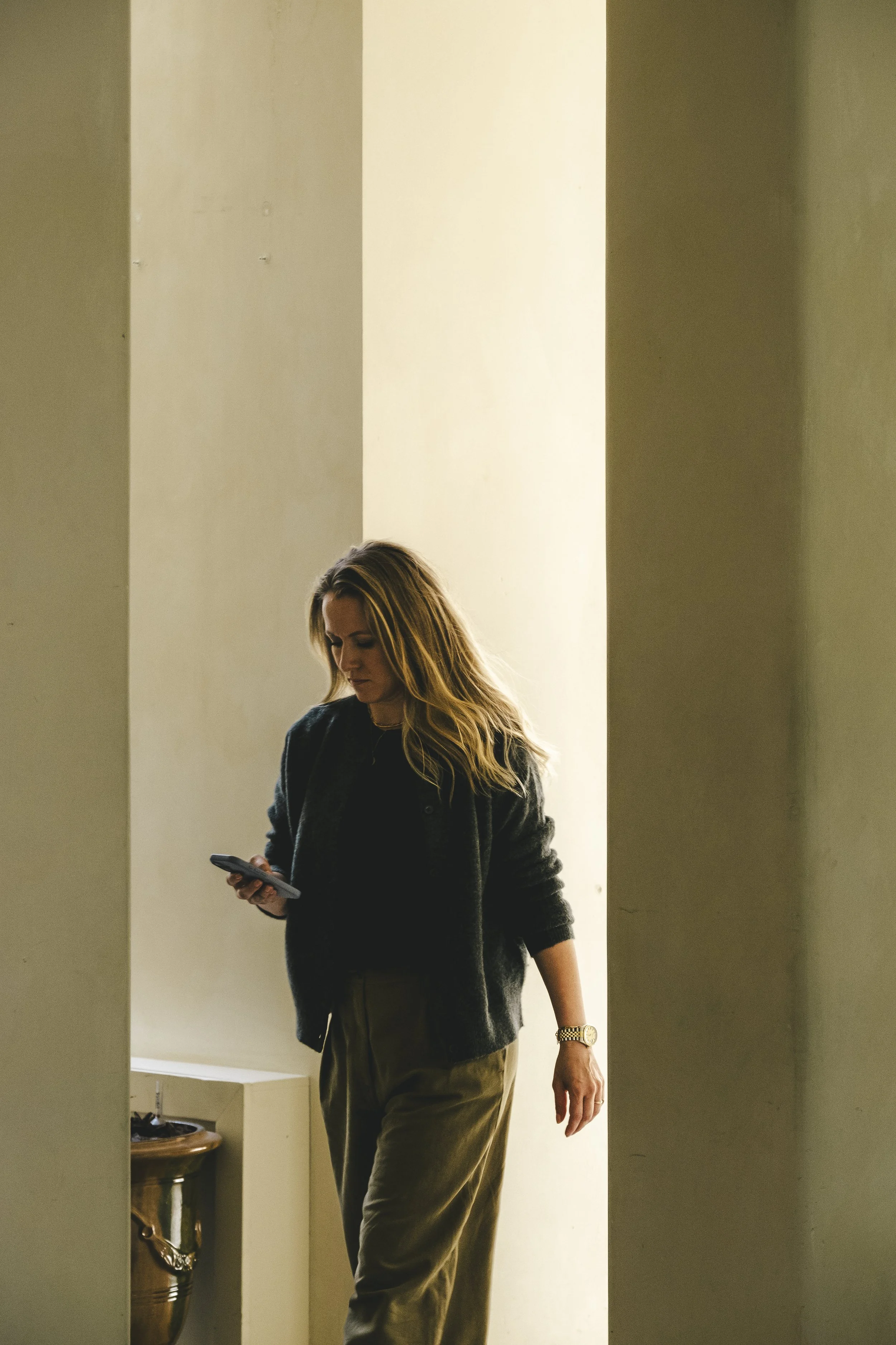 A woman with long blonde hair wears a dark jacket and olive-green pants, standing and looking at her phone in a room with beige walls and a small table on the left.