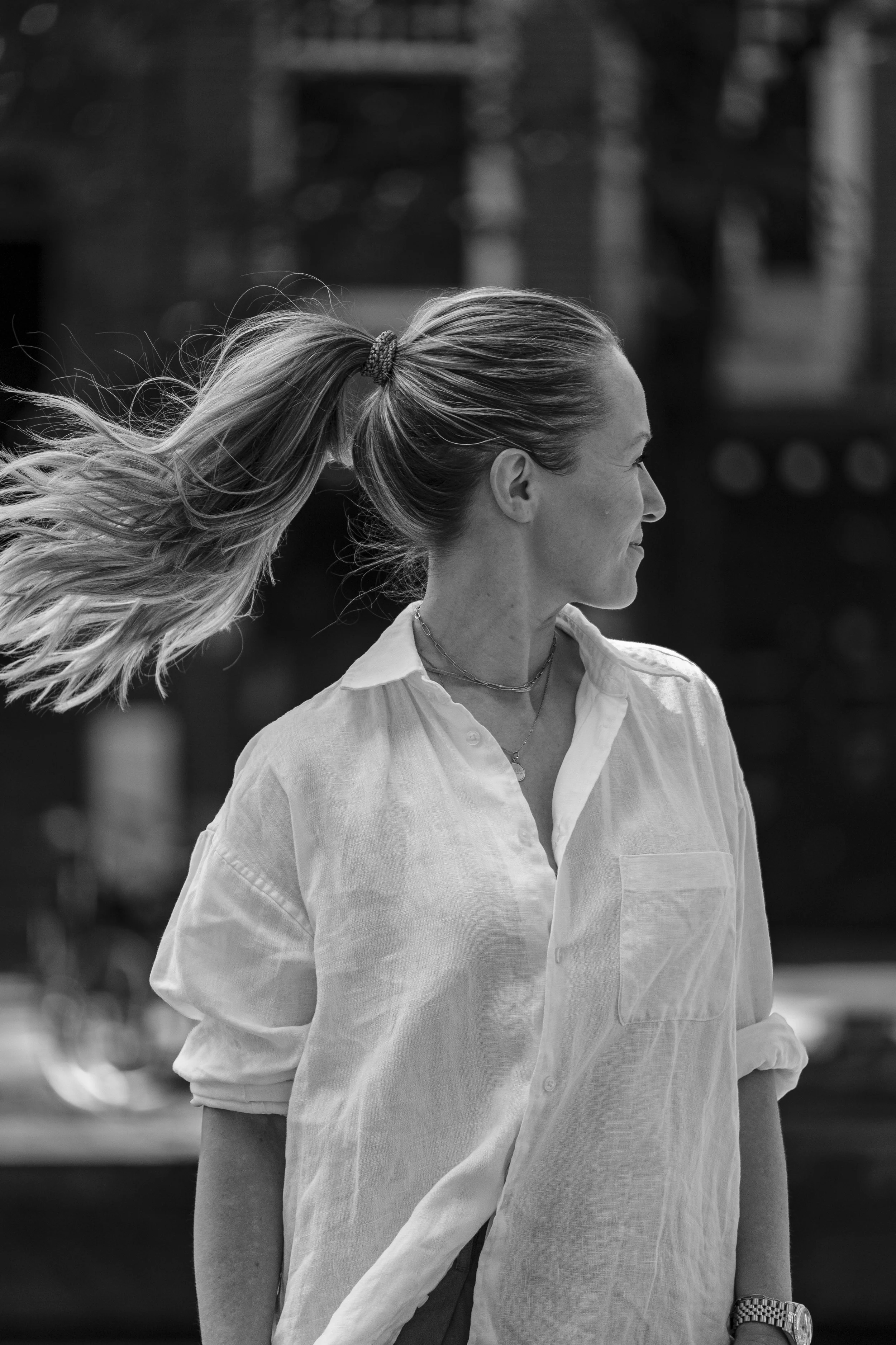 A woman with long hair tied back in a ponytail, wearing a light-colored shirt with rolled-up sleeves, standing outdoors during daytime.