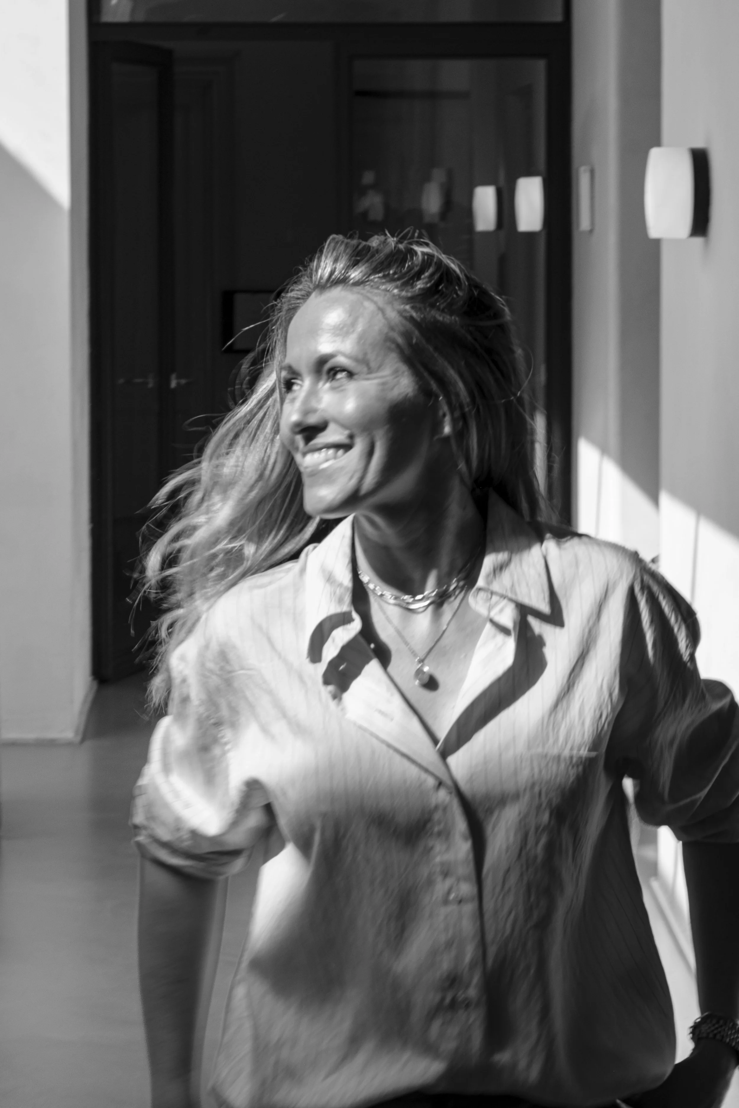 Black and white photo of a woman smiling with long hair, wearing a button-up shirt, standing in a room with a door and wall-mounted objects.