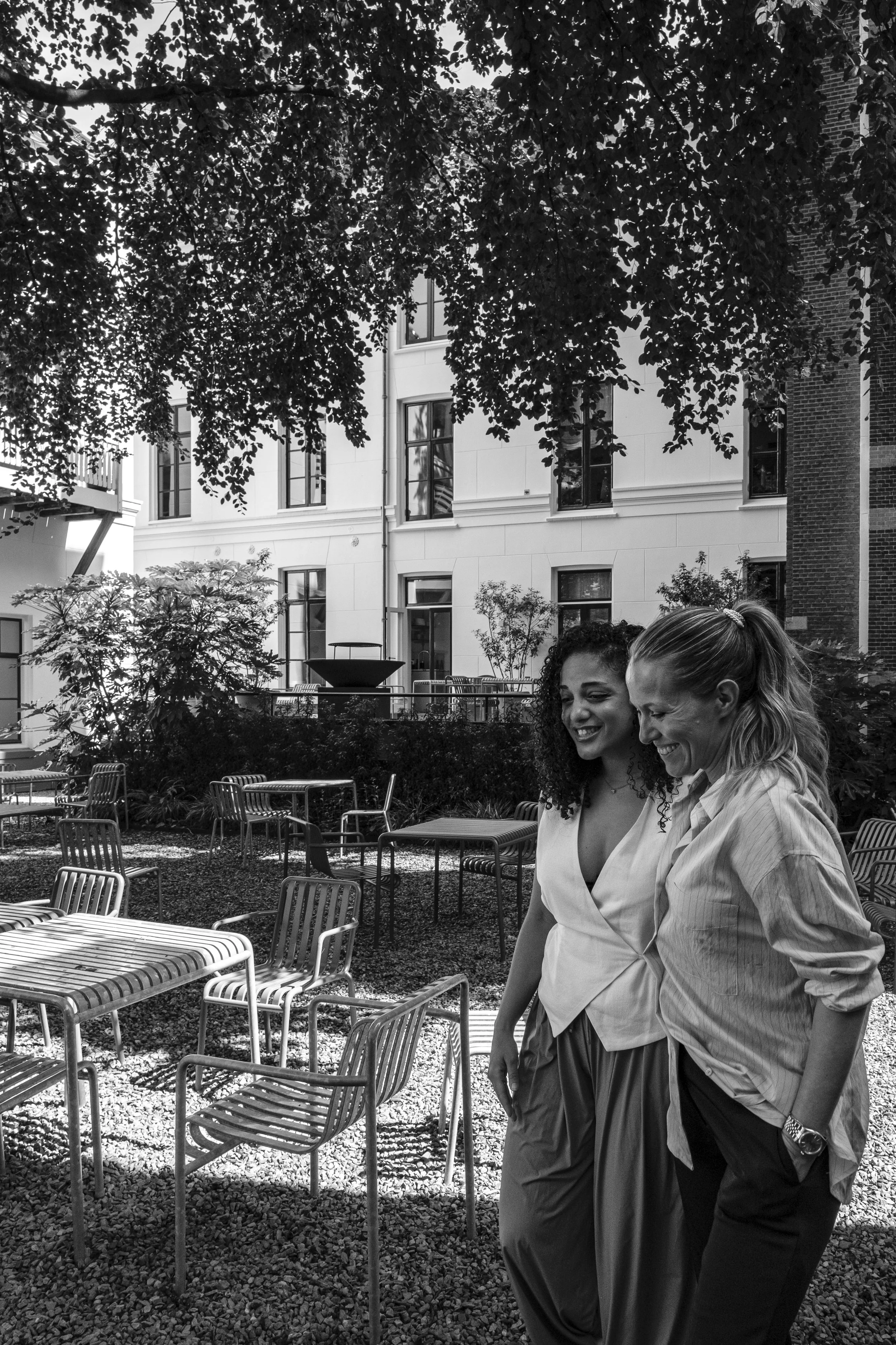 Two smiling women walking outdoors in a garden or patio area, with tables and chairs, surrounded by trees and building facades.