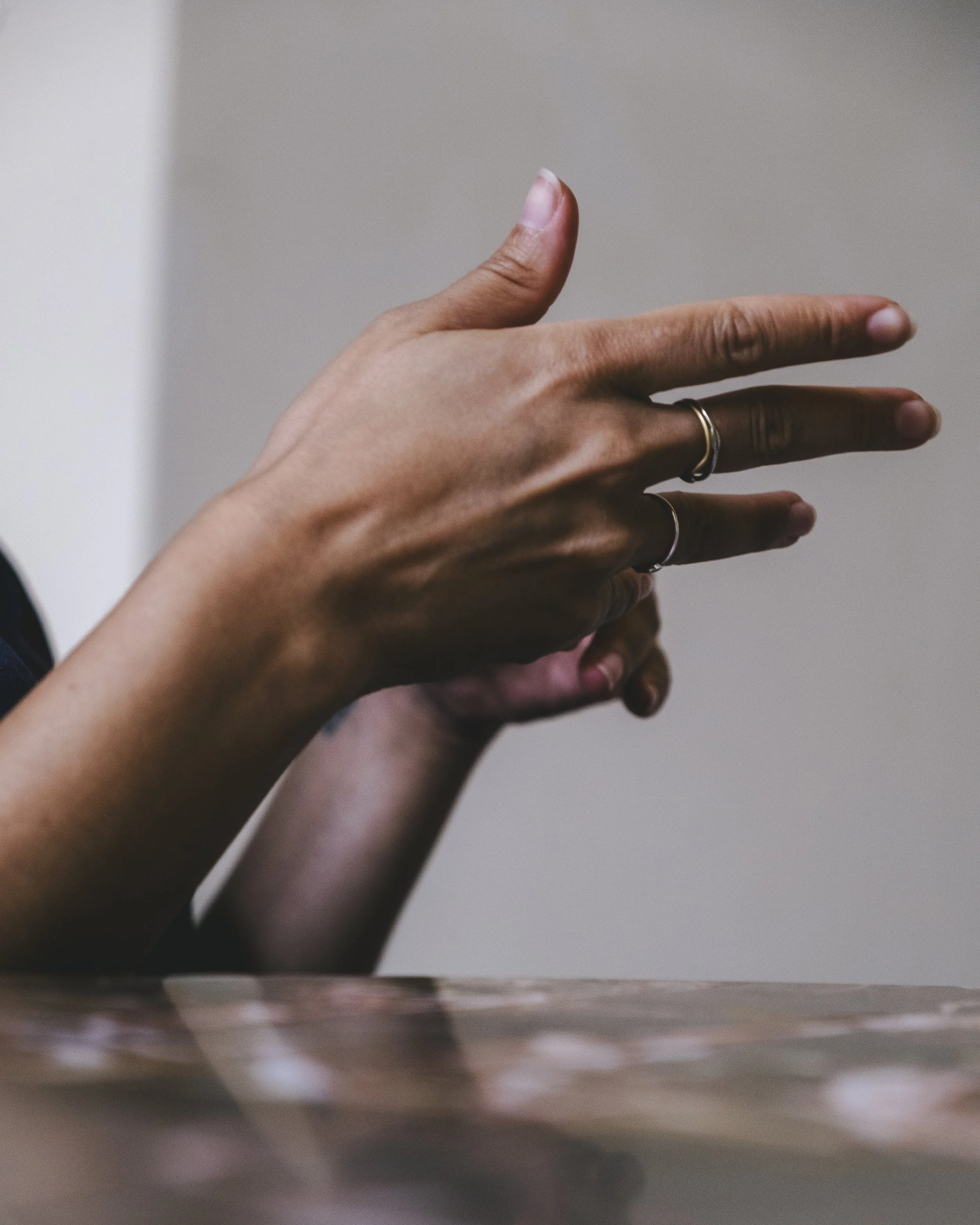 Close-up of a person's hand with multiple rings, making a peace sign gesture, seen from below against a plain background.
