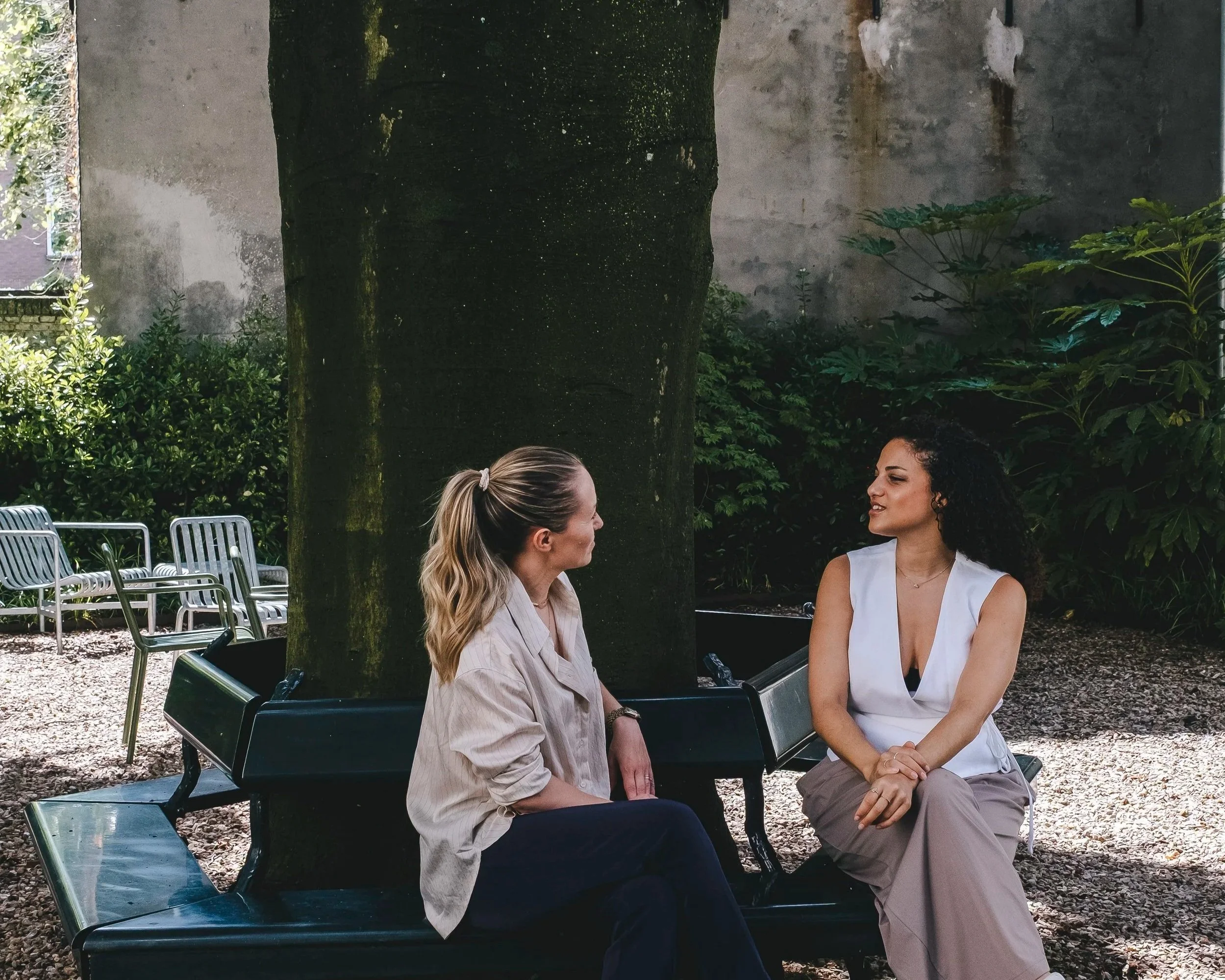 Two women sitting on a park bench engaged in conversation. One woman has light hair tied in a ponytail and is wearing a beige jacket, the other has dark curly hair and is wearing a white sleeveless top. There are empty park chairs and a large tree in the background, with greenery and a concrete wall.