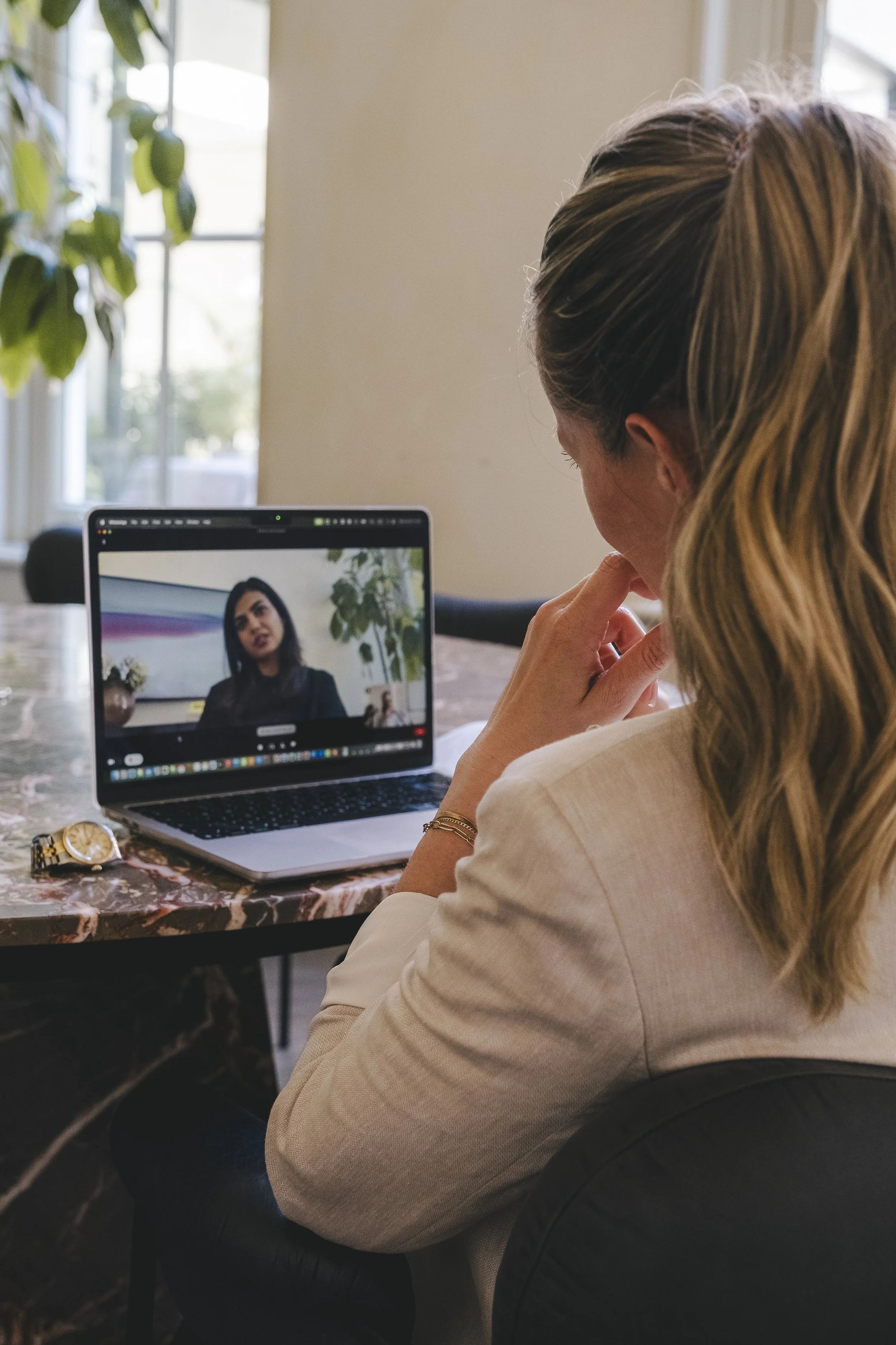Woman with blonde hair in a ponytail sitting at a table, participating in a video call on her laptop, with another woman visible on the screen.