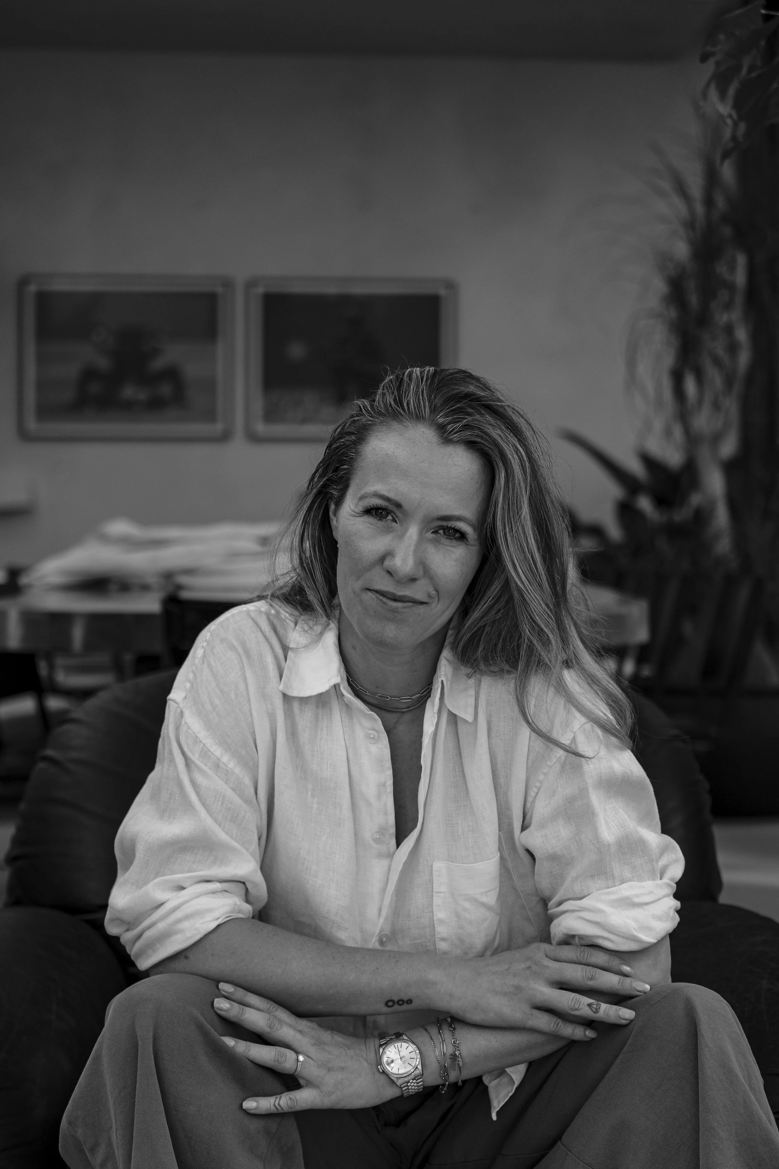 A woman with wavy hair and light skin sitting on a comfortable chair in an indoor setting, smiling and looking at the camera. She is wearing a white shirt with rolled-up sleeves, a watch, and bracelets. In the background, there are framed pictures on the wall and some plants.