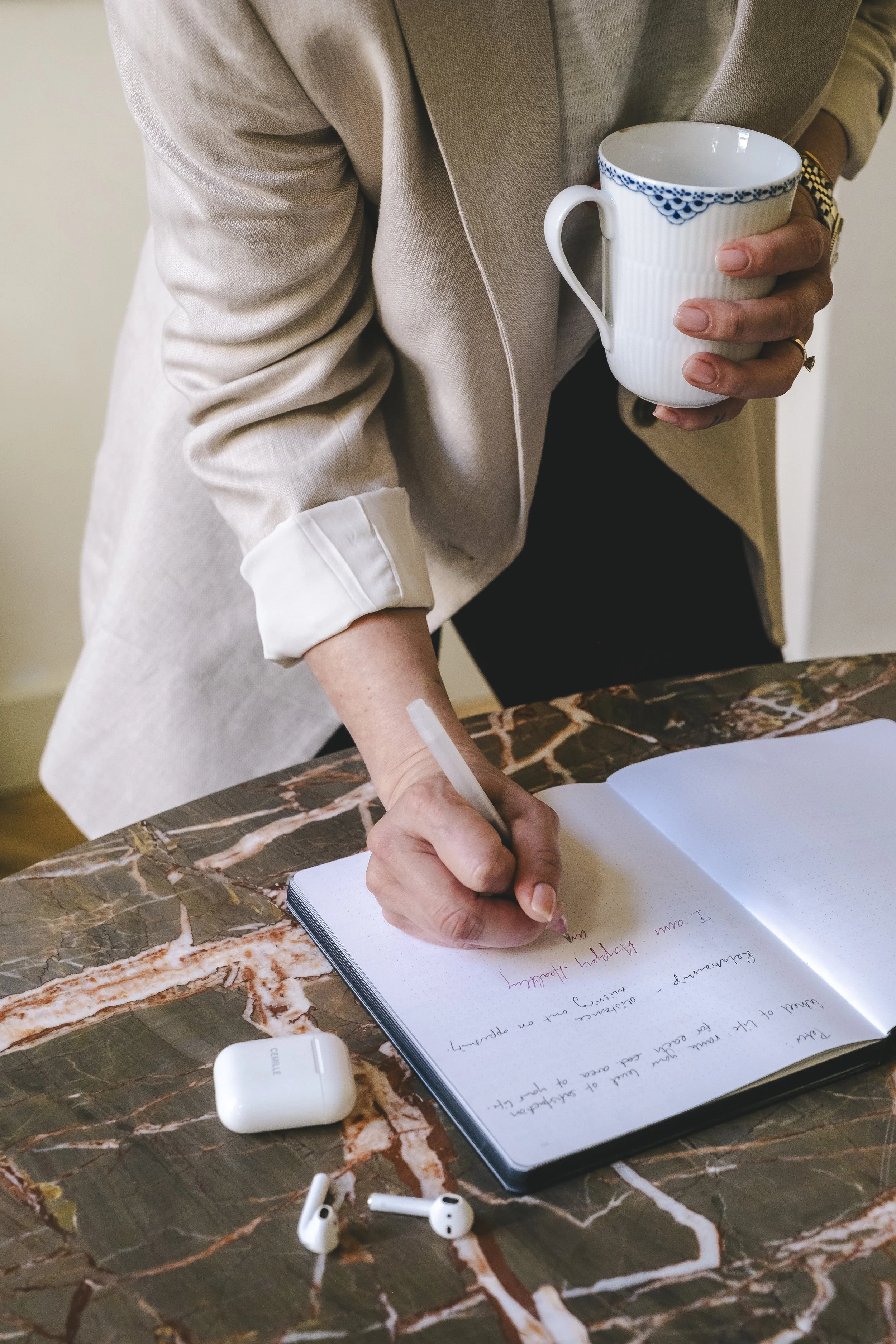 Person wearing a light beige blazer writing in a notebook on a marble table, holding a white mug with blue patterns, and AirPods resting on the table.