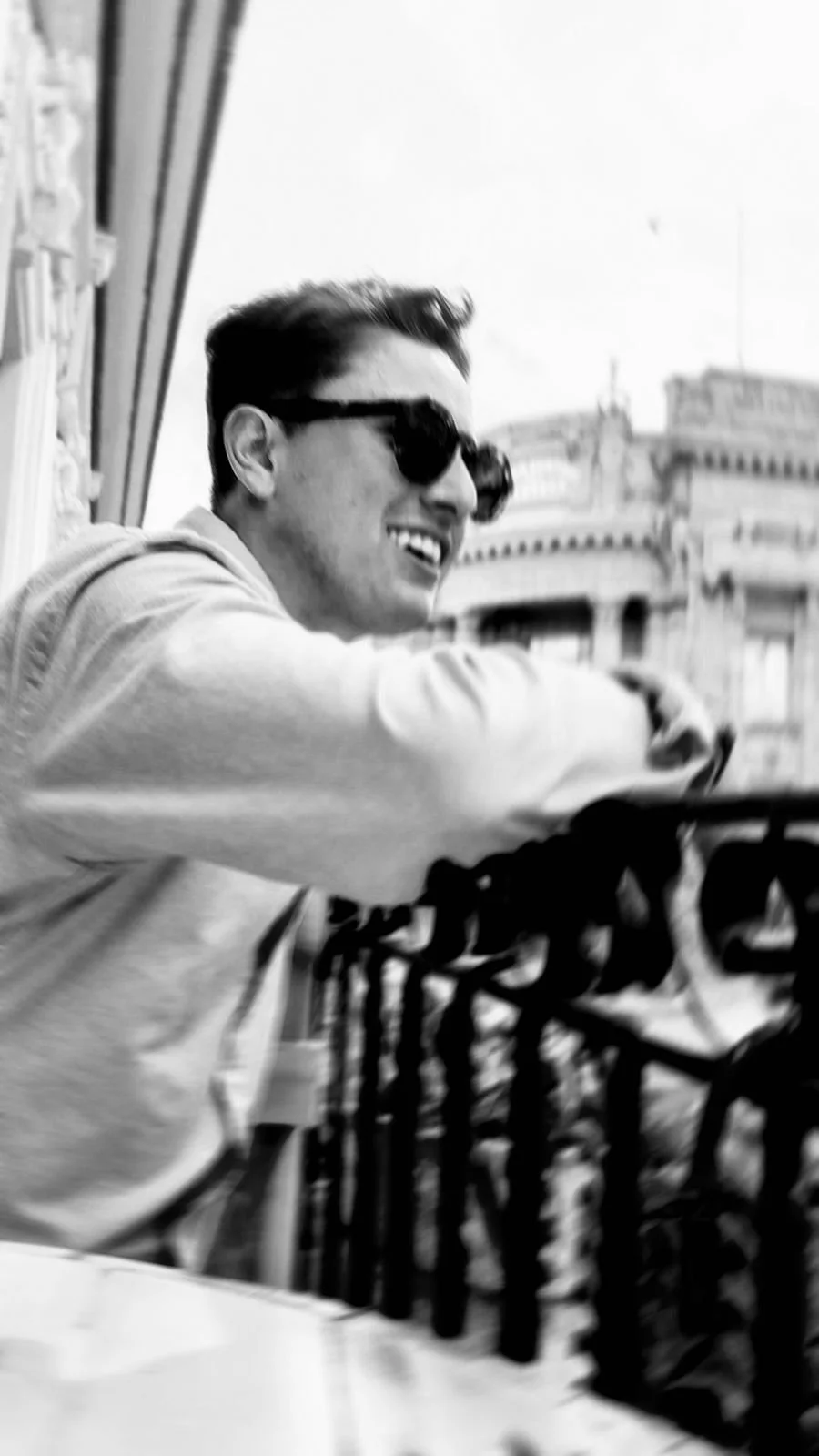 Joven sonriendo con gafas de sol, en un entorno urbano en blanco y negro.