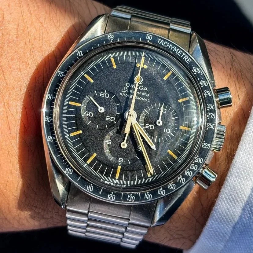 Reloj Omega Speedmaster Professional en la muñeca, con esfera negra, agujas doradas y pulsera de metal.
