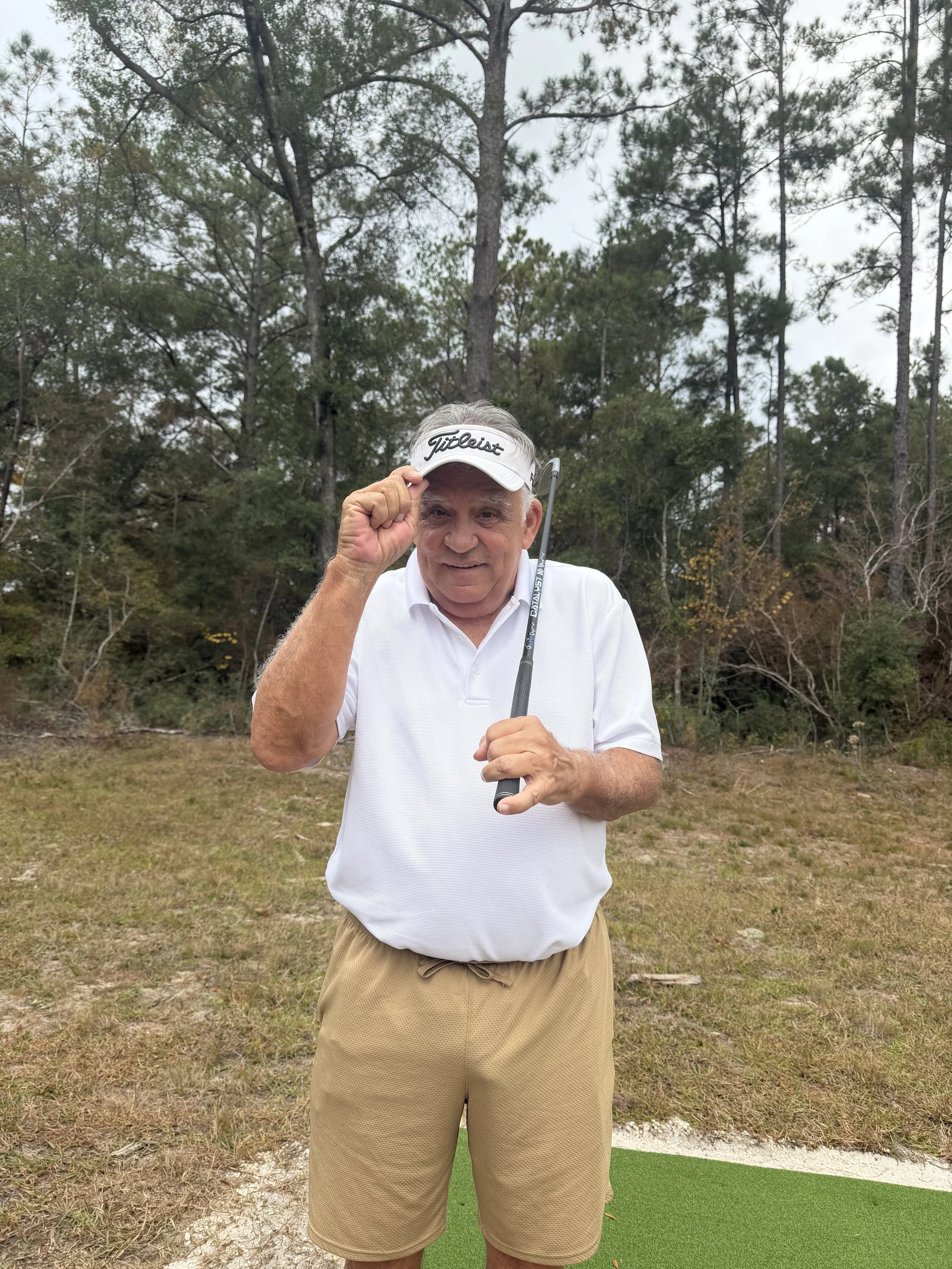 An elderly man smiling outdoors on a cloudy day, wearing a white polo shirt, khaki shorts, a Titleist visor, and holding a golf club, with a forested background and a small green mat under his feet.