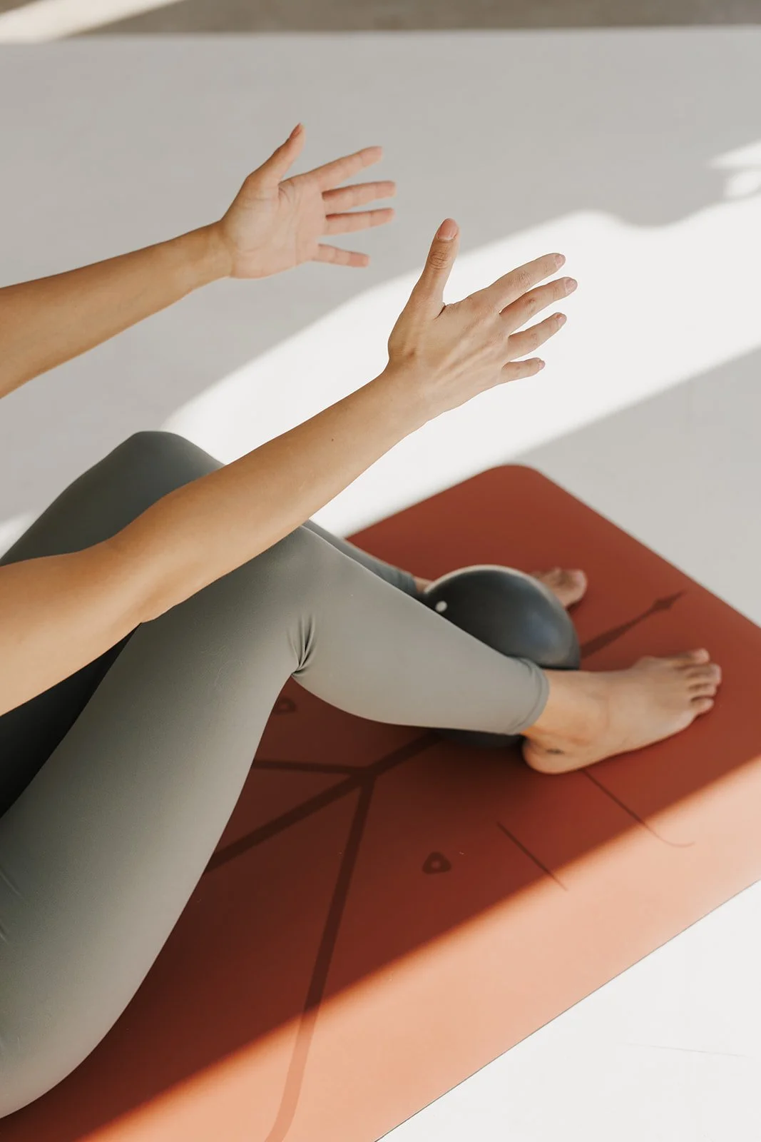 Person doing yoga on a red mat with a black massage ball, sitting cross-legged, stretching their arms upward, in a bright room.