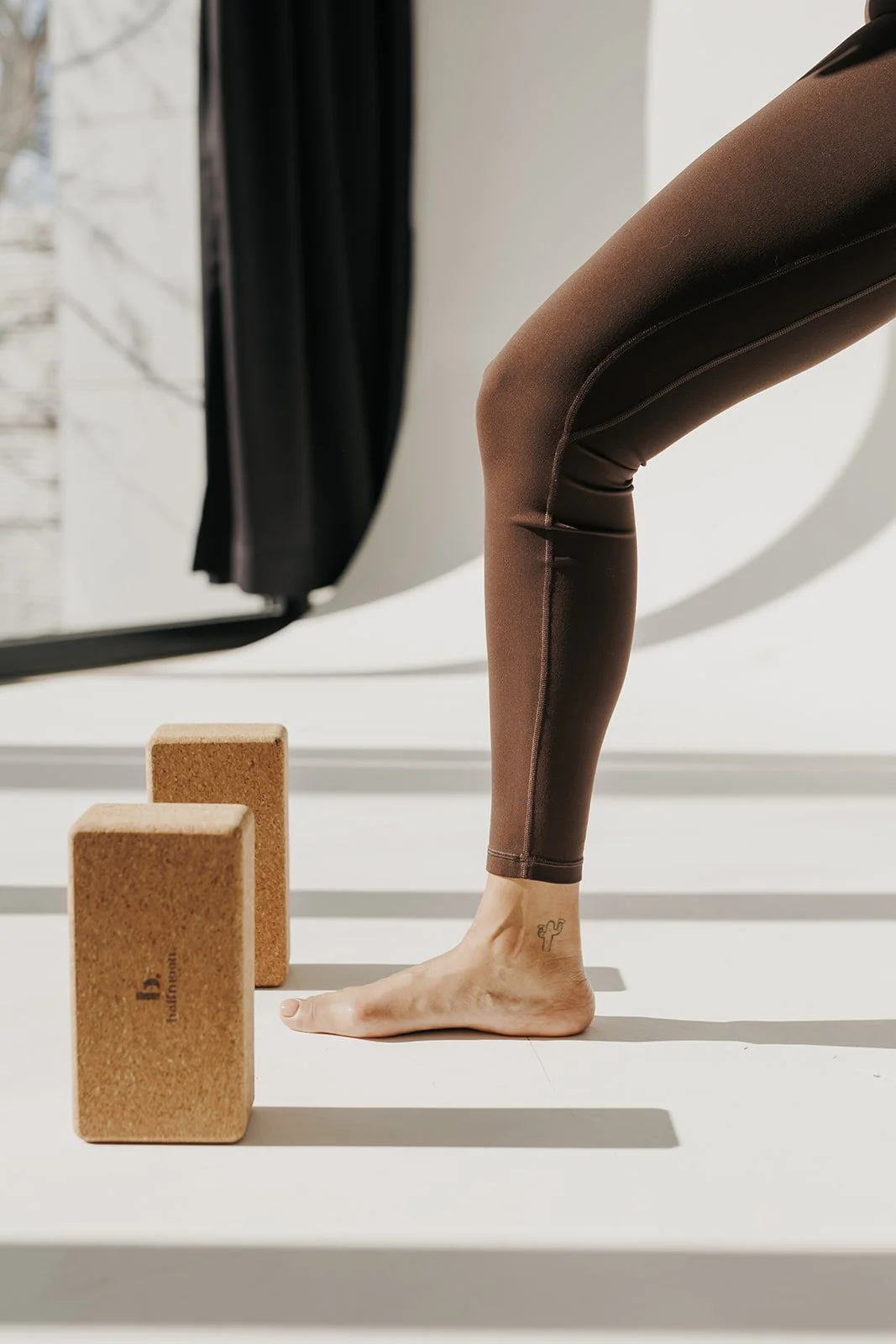 A person doing yoga or stretching, with one foot flat on the ground and the other knee bent, on a white surface near a window with black curtains. Two cork yoga blocks are nearby.