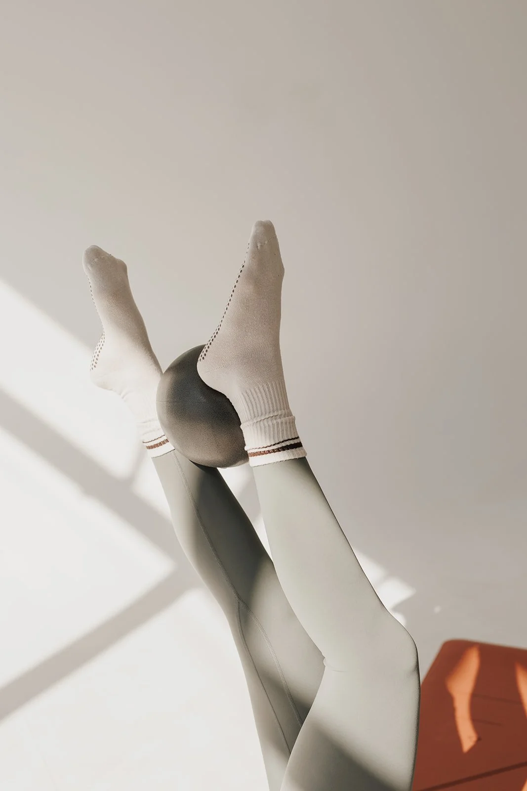 Two legs in gray leggings and white socks holding a black exercise ball against a plain wall with sunlight and shadow.