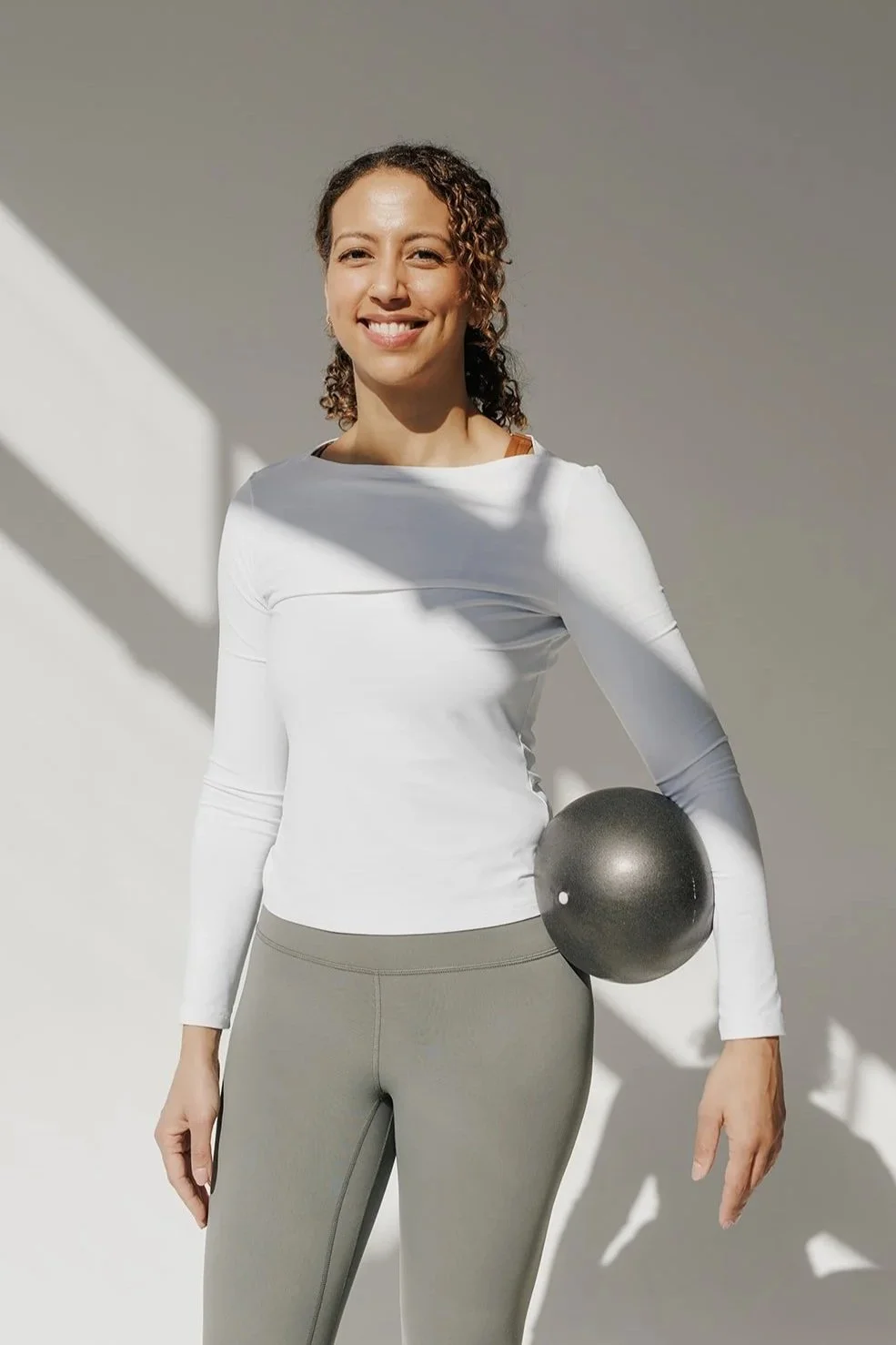 A woman with curly hair smiling and holding a black exercise ball in her right hand, standing against a plain light-colored background with shadows.