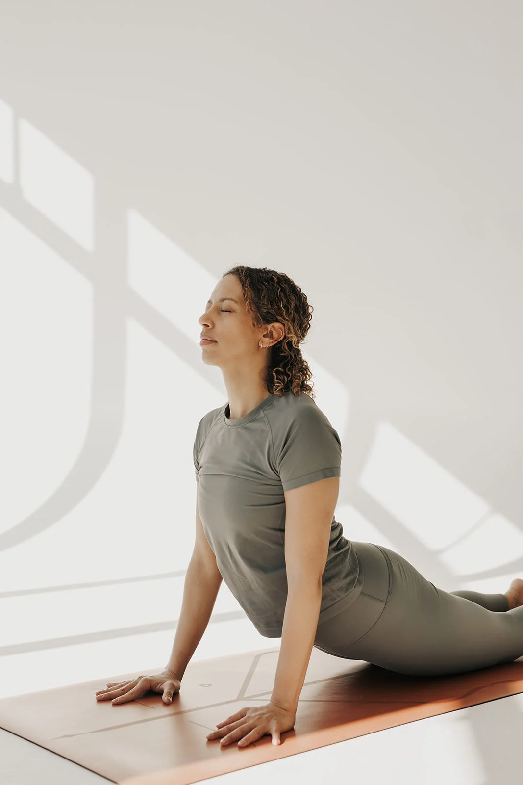 Woman practicing yoga in a sunlit room, in a cobra pose with eyes closed and a peaceful expression.