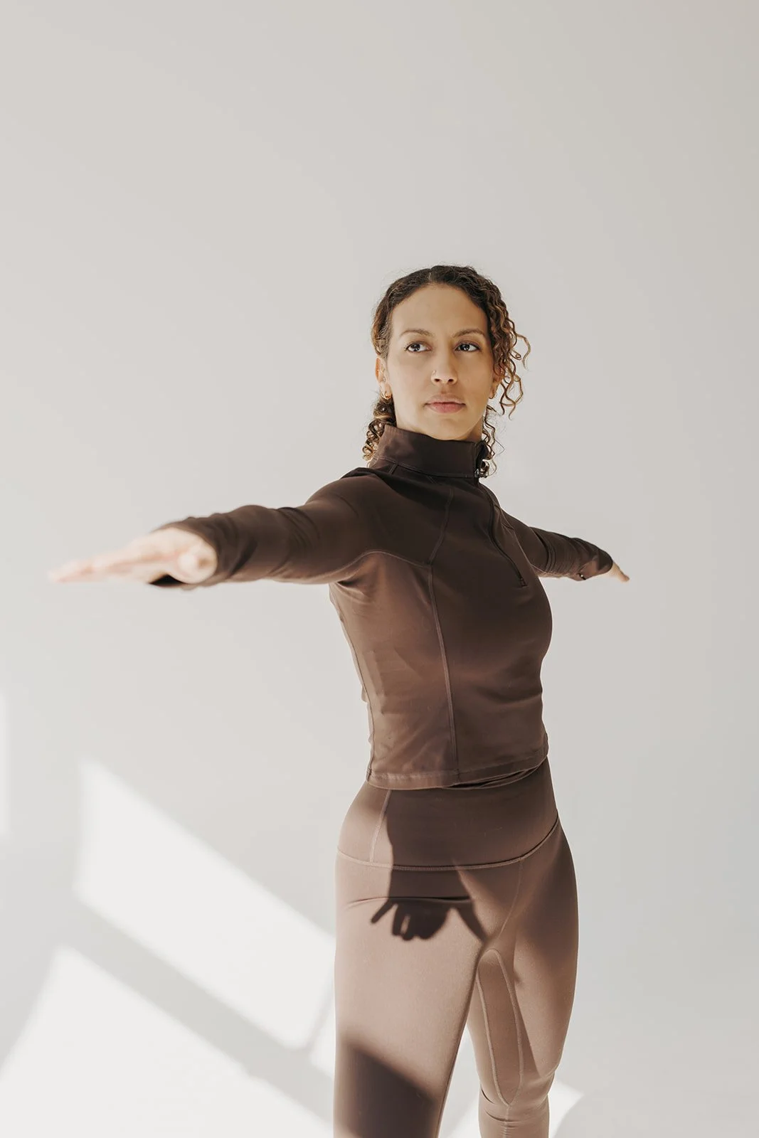 A woman in workout attire standing with arms extended horizontally, facing forward in a bright, minimalist setting.