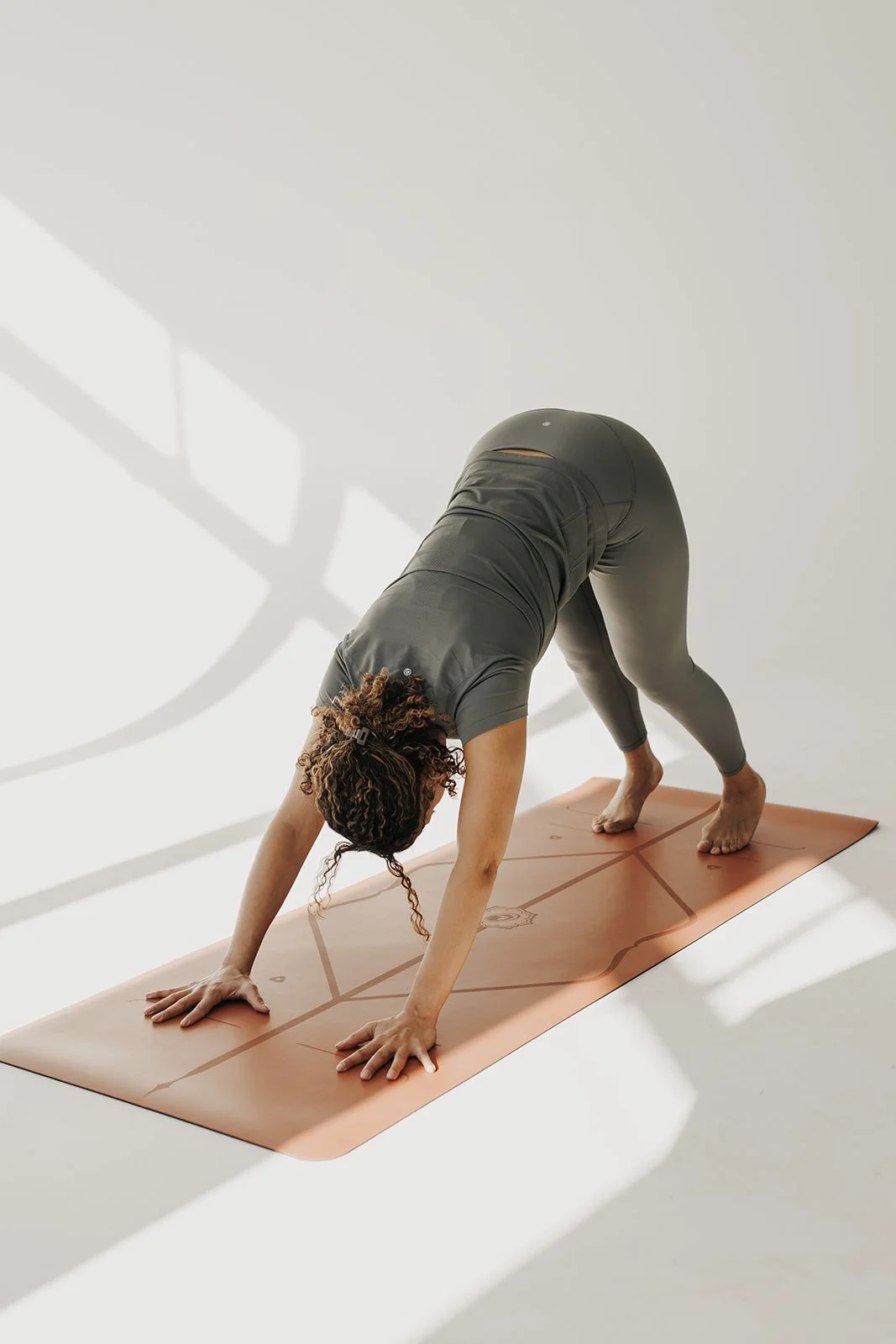 A woman in black athletic wear practicing yoga in downward dog pose on a pink yoga mat in a bright, minimally decorated room.