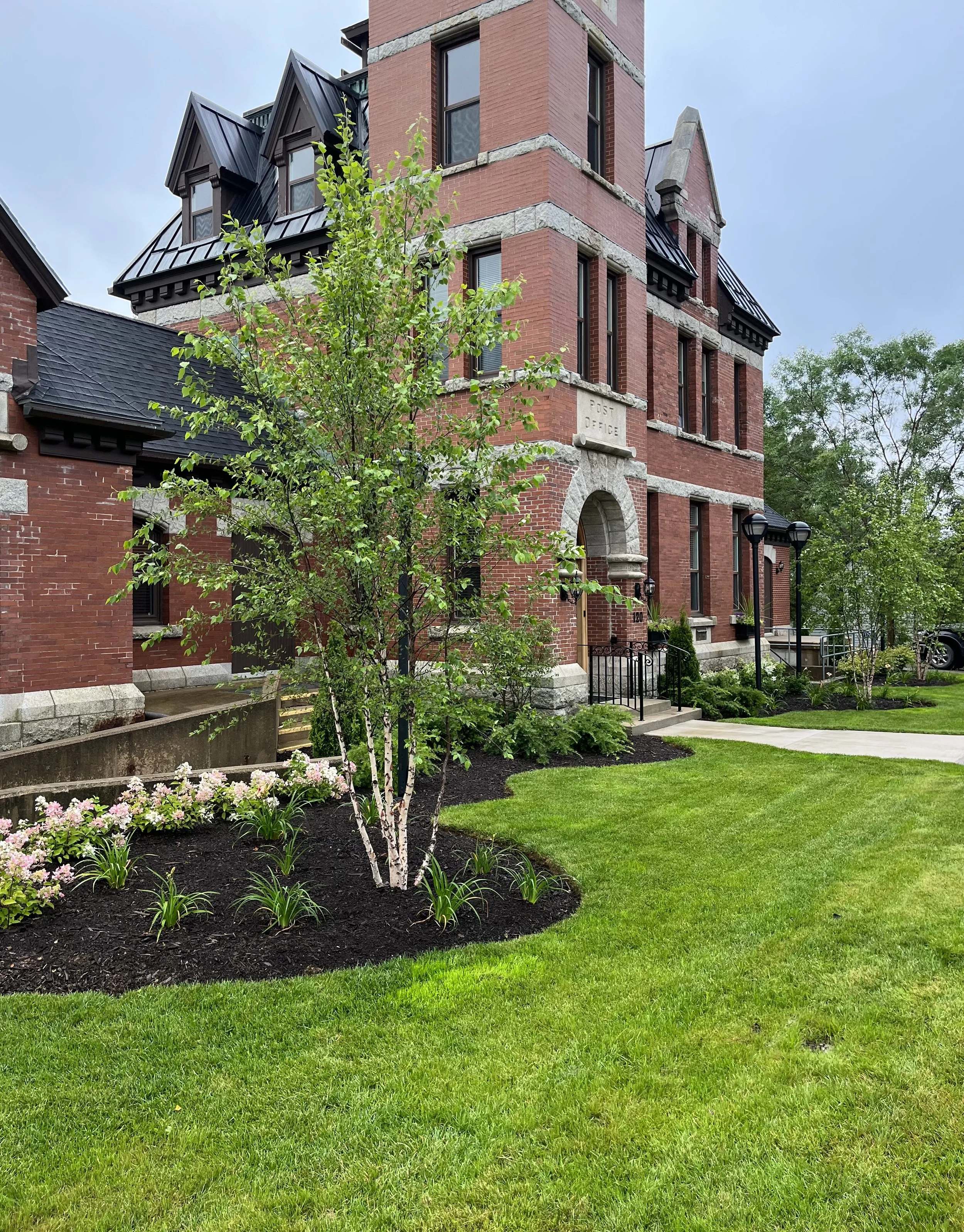 A brick building with a small landscaped garden, young trees, flowering plants, and a sidewalk in front.