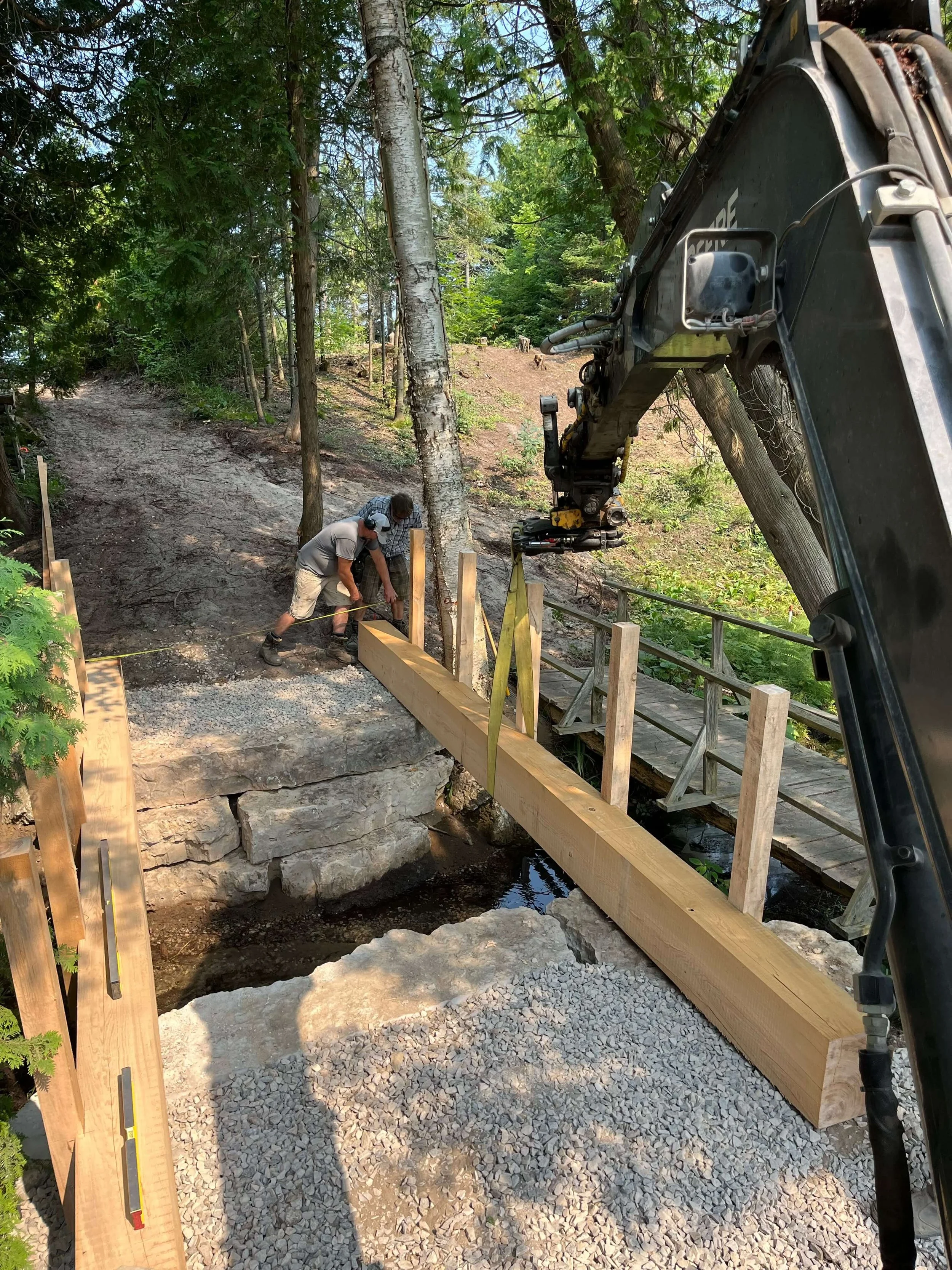 A construction site in a wooded area with workers building a small bridge over a creek. There is a large excavator with its arm extended, and wooden beams forming the sides of the bridge. The ground is covered with gravel and dirt, and trees surround the scene.