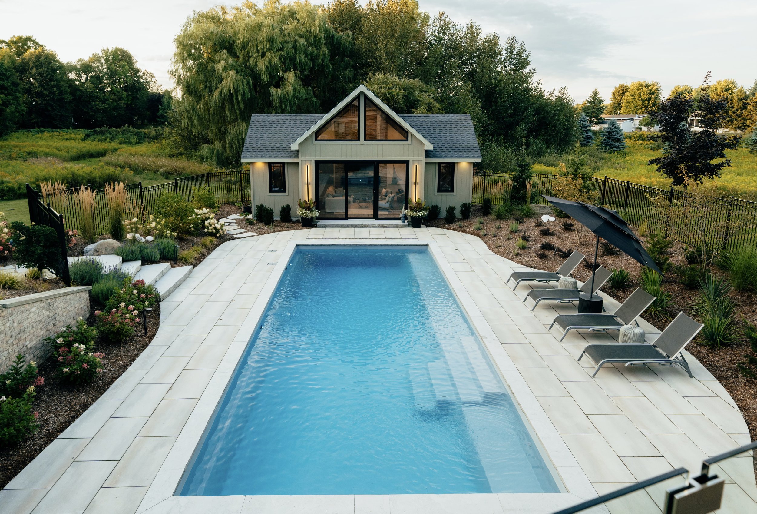 A backyard with a rectangular swimming pool surrounded by white tiles, lounge chairs, an umbrella, landscaped plants, and a modern house with large glass doors and windows, all enclosed by a black metal fence and lush green trees.