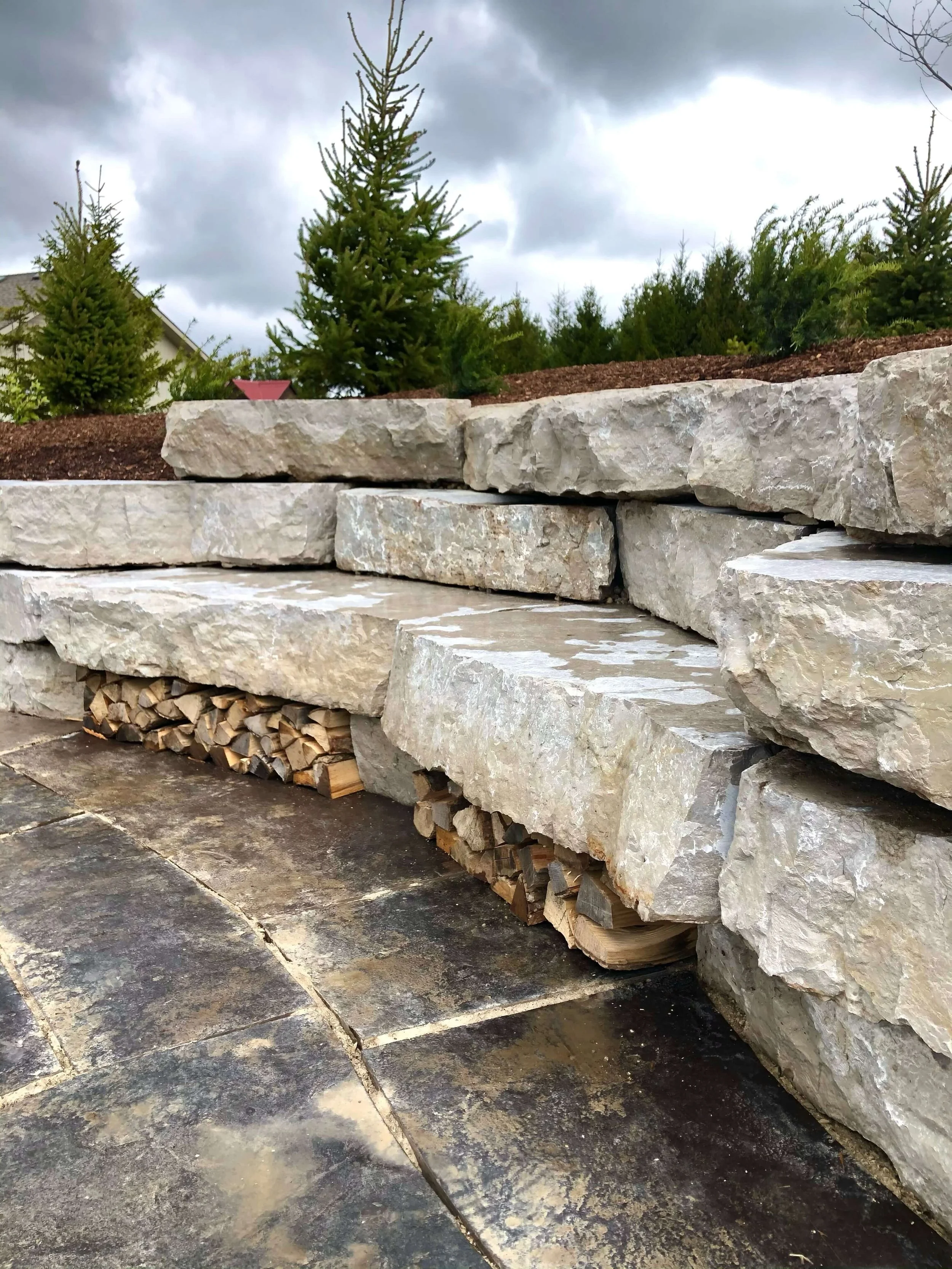 Stacked large natural stone slabs with firewood underneath on a cloudy outdoor setting.