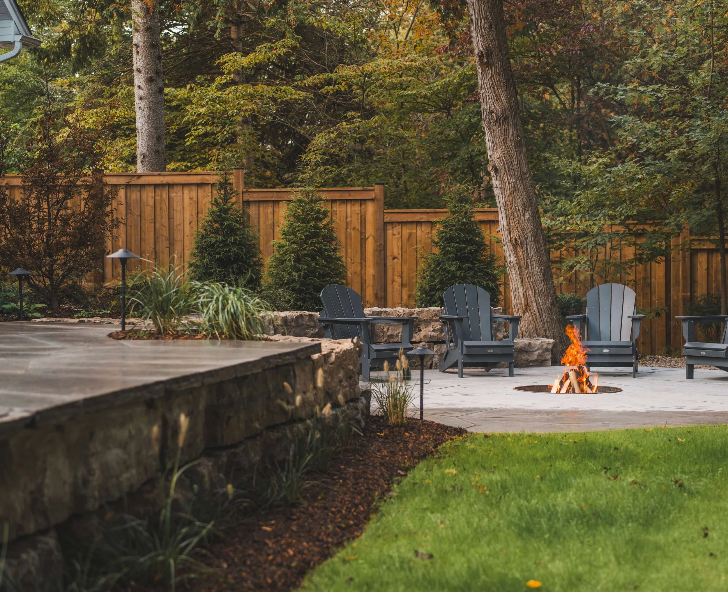 A backyard with a wooden fence, tall trees, small evergreen bushes, a fire pit with a small bonfire, Adirondack chairs, and landscape lighting during daytime.