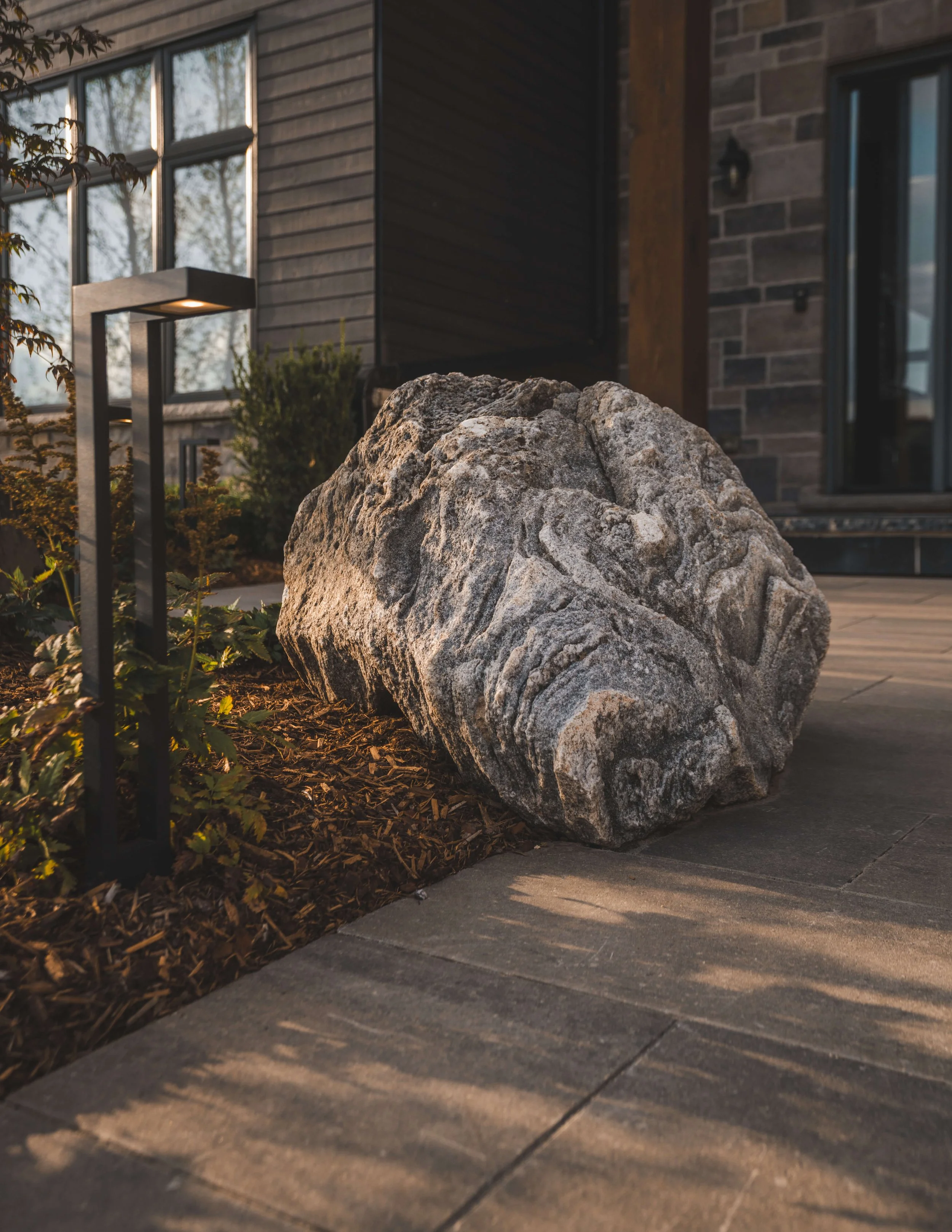 Large gray rock in front of a modern house with wood and brick exterior, illuminated by outdoor lights with plants and a sidewalk.