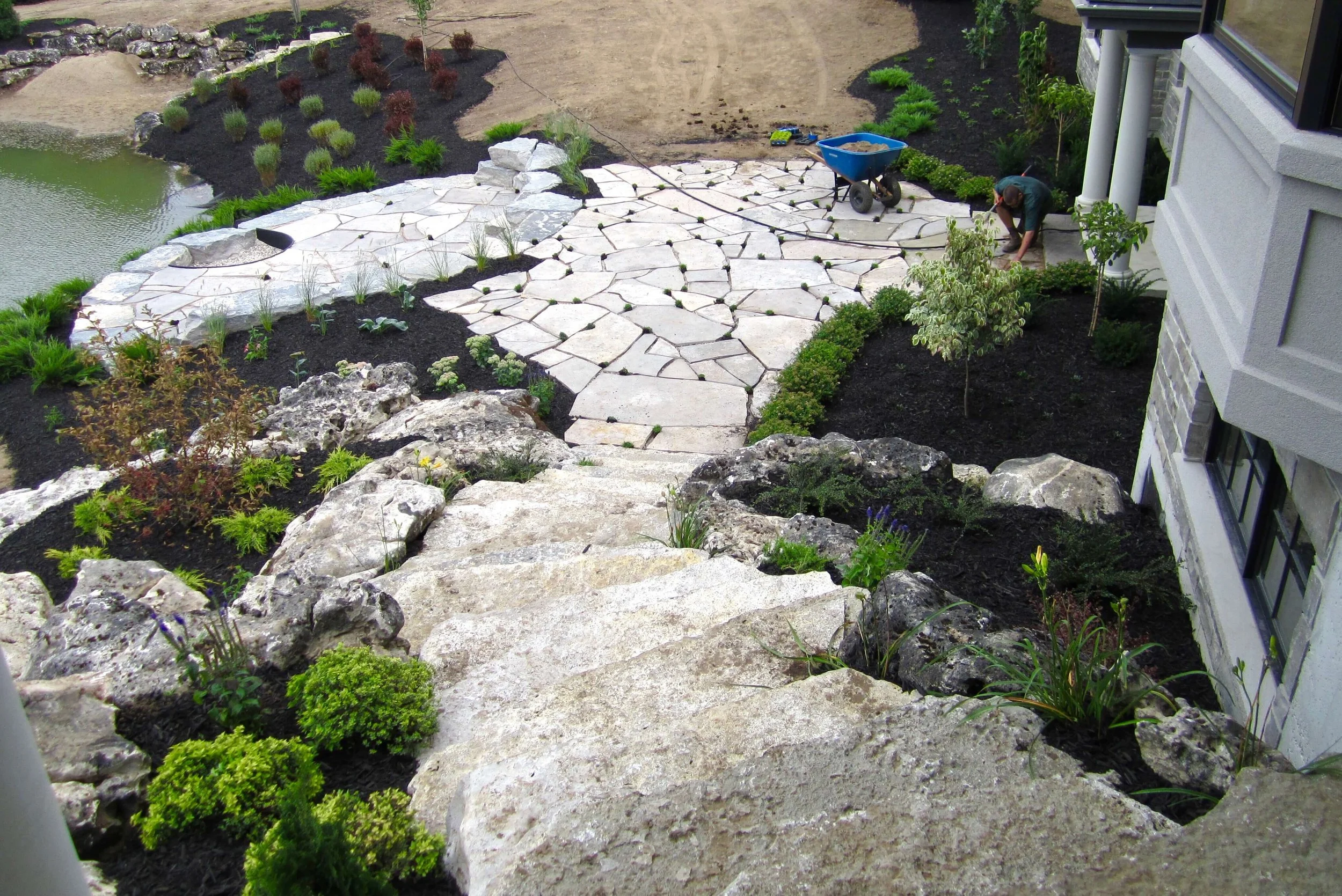 Construction worker laying large stone slabs on a patio next to a house with plants around.