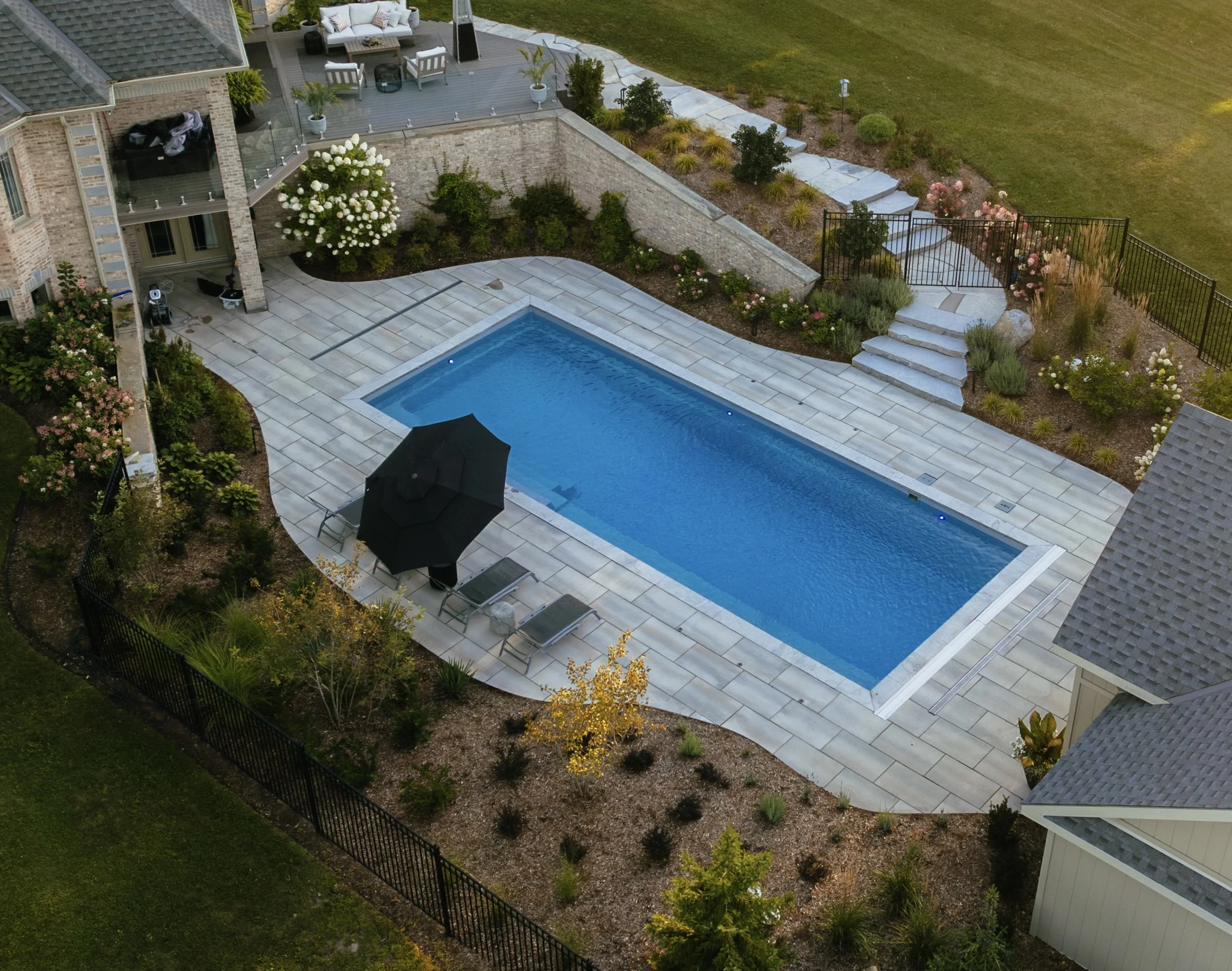 A backyard with a swimming pool, a patio with outdoor furniture, a garden with plants, shrubs, and flowers, and a staircase leading up to an elevated deck area.