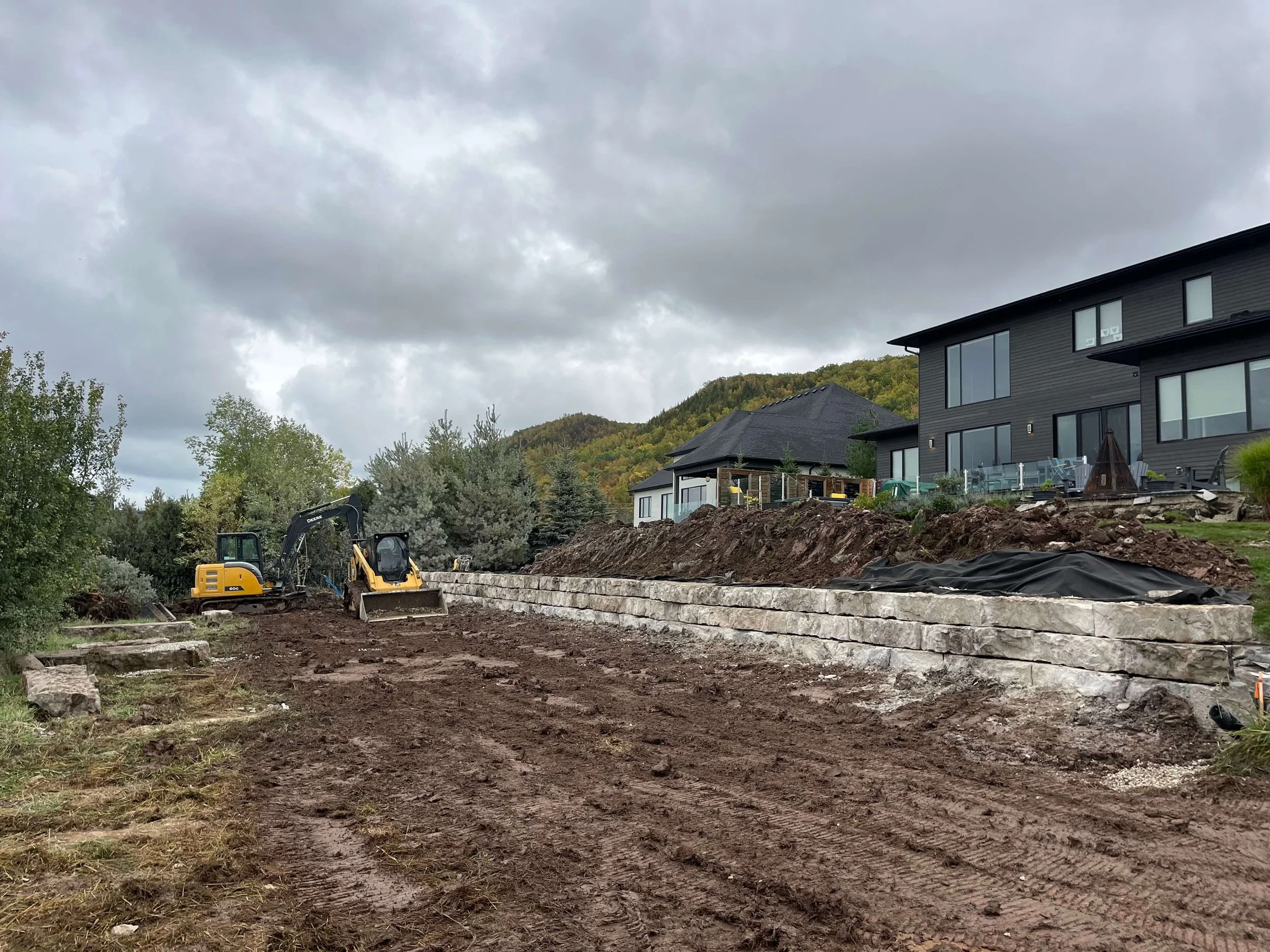 Construction site with small excavator and house in the background, with a cloudy sky.