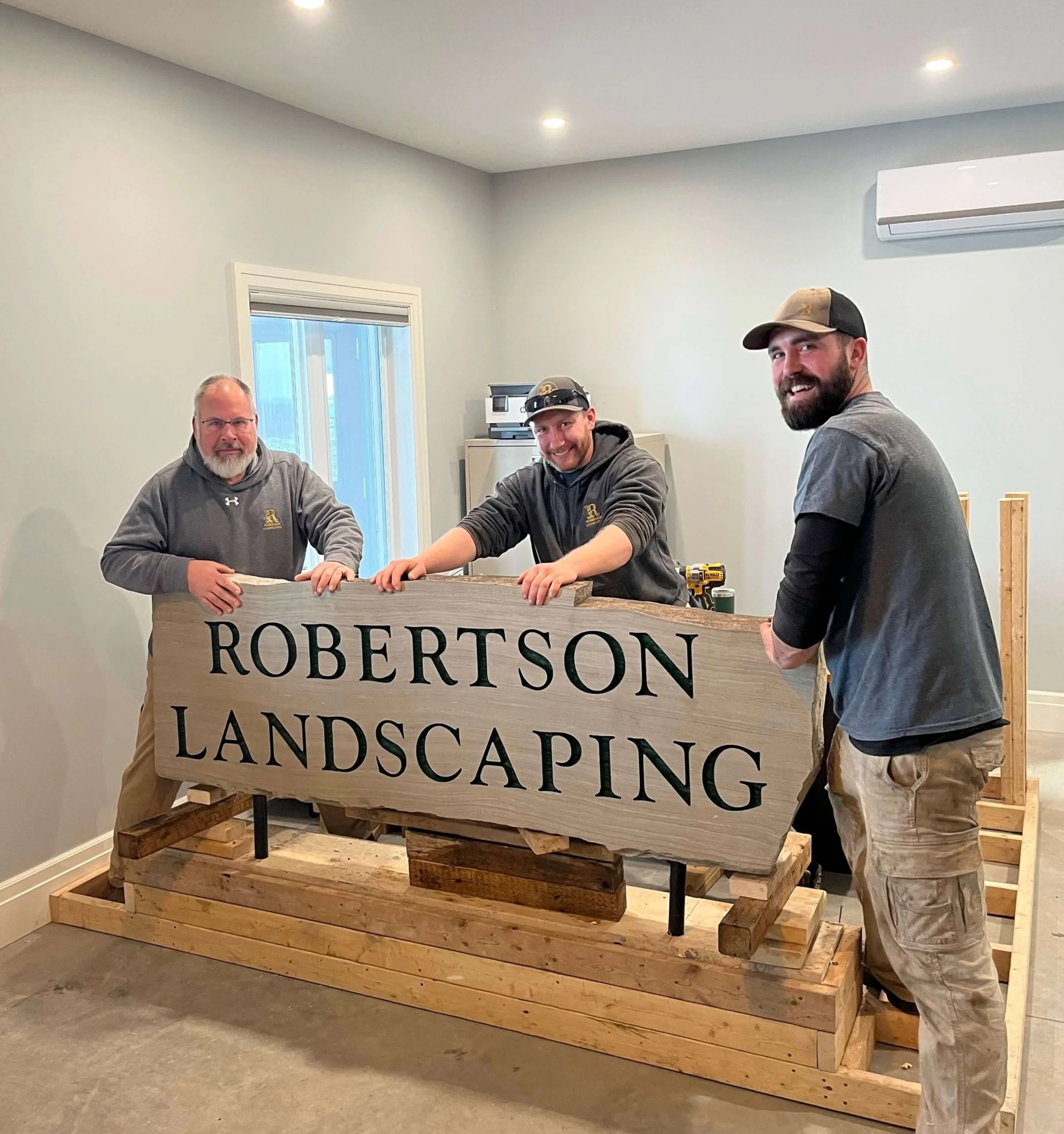 Three men working on a wooden landscaping sign that reads 'ROBERTSON LANDSCAPING' in an indoor space with gray walls and ceiling, a small window, and an air conditioning unit.