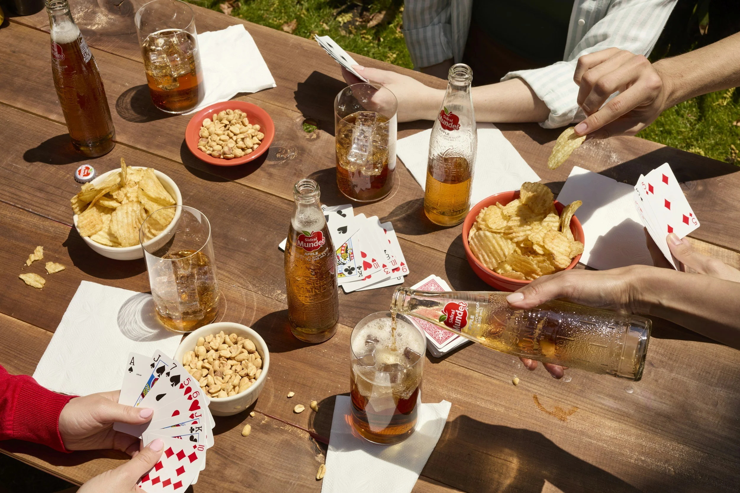 A group of people playing cards at an outdoor wooden table, with snacks like potato chips, peanuts, and bottles of soda. Some hands are visible holding cards or placing chips on the table, and several glasses of soda are on the table.