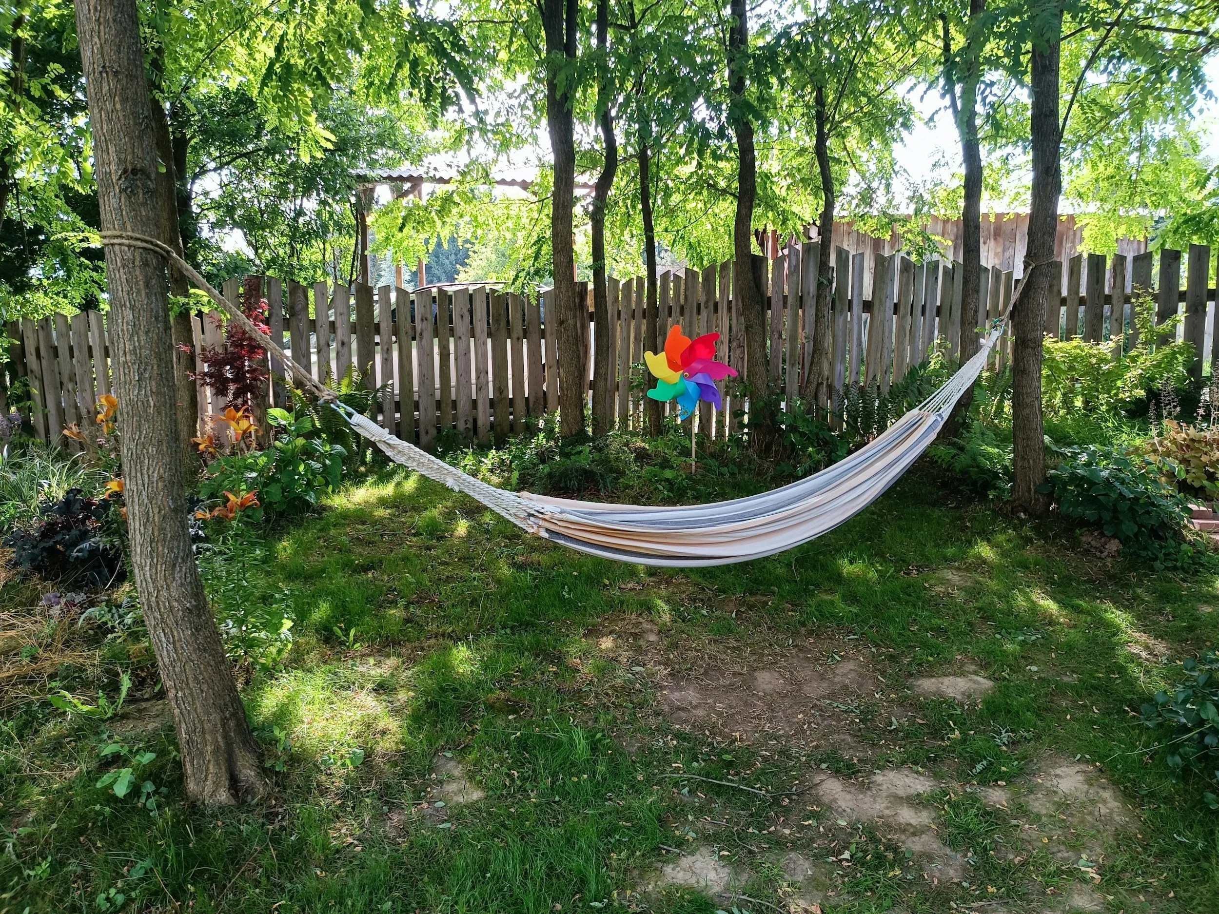 A backyard with trees, a wooden fence, a hammock, and a colorful wind spinner in the garden.
