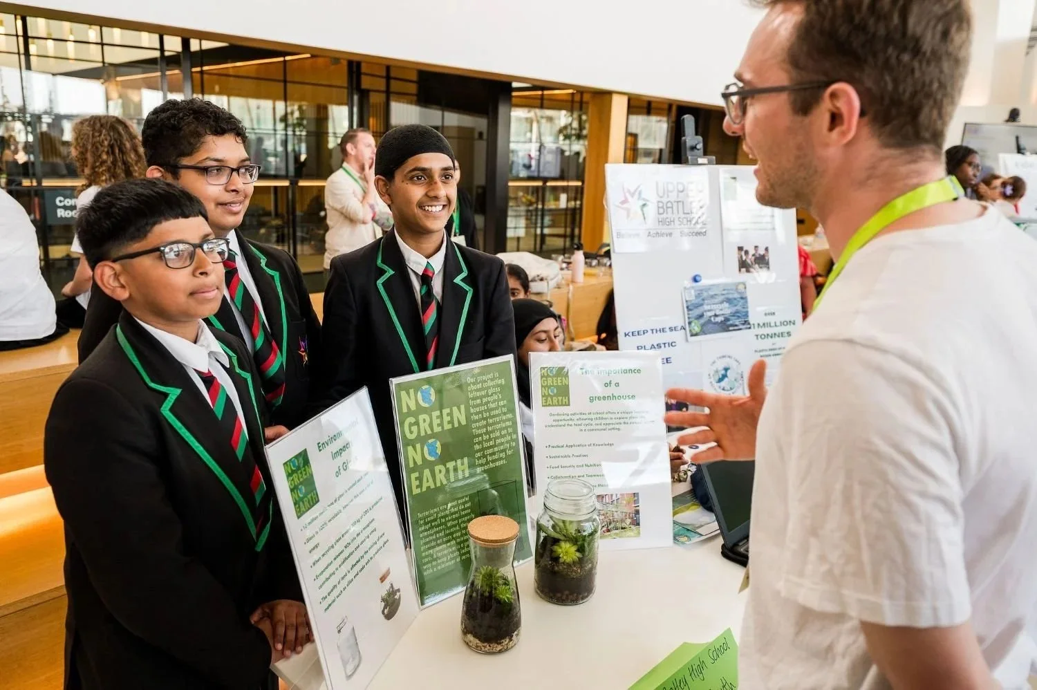 A group of school children in black uniforms with green trim and striped red, black, and green ties are talking to a man at a science fair. The children are standing in front of display boards about environmental sustainability and recycling, with glass jars containing plants on the table.