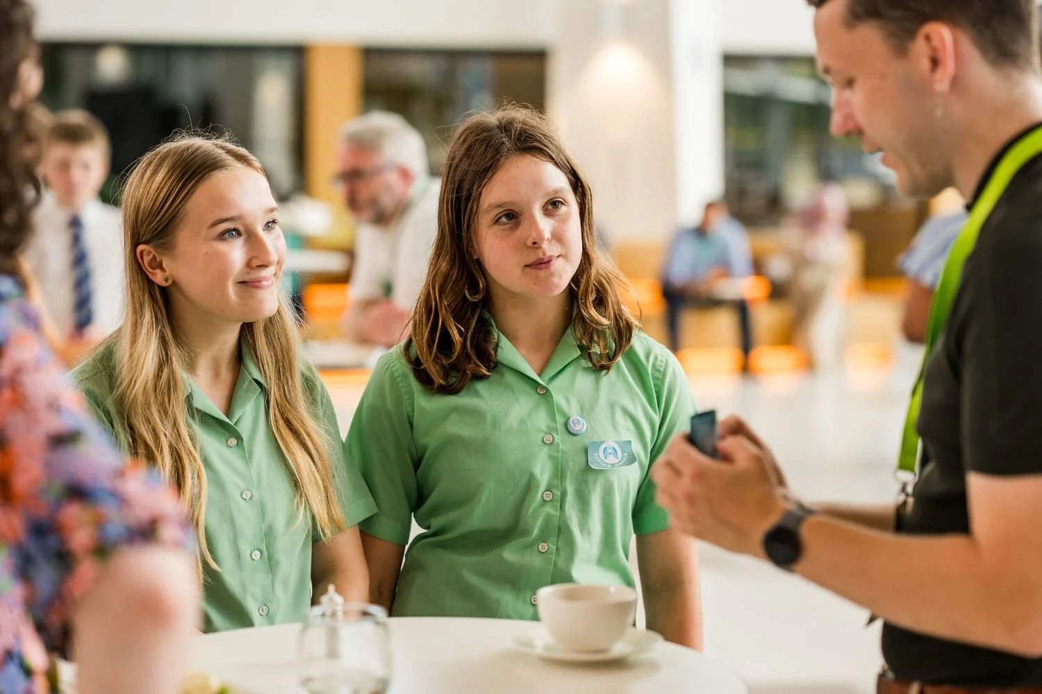 Two young girls in green uniforms talking to a man at a restaurant or cafe. The girls are listening attentively, and the man is holding a small object, possibly a phone, while explaining something.