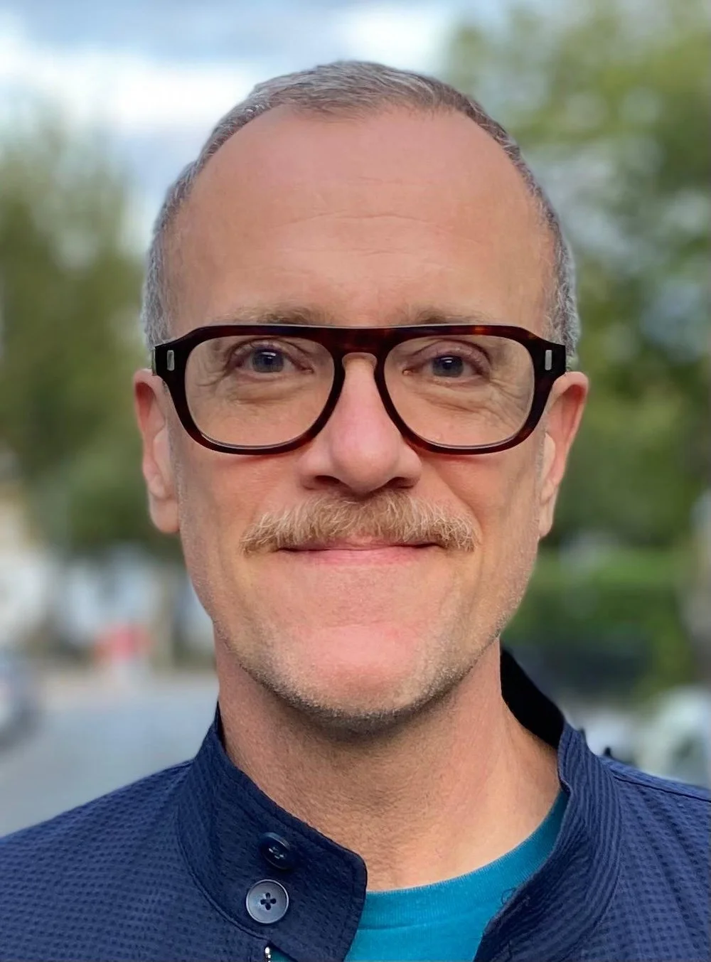 Close-up of a man with glasses, light beard, mustache, and short hair, smiling outdoors with blurred green trees in the background.