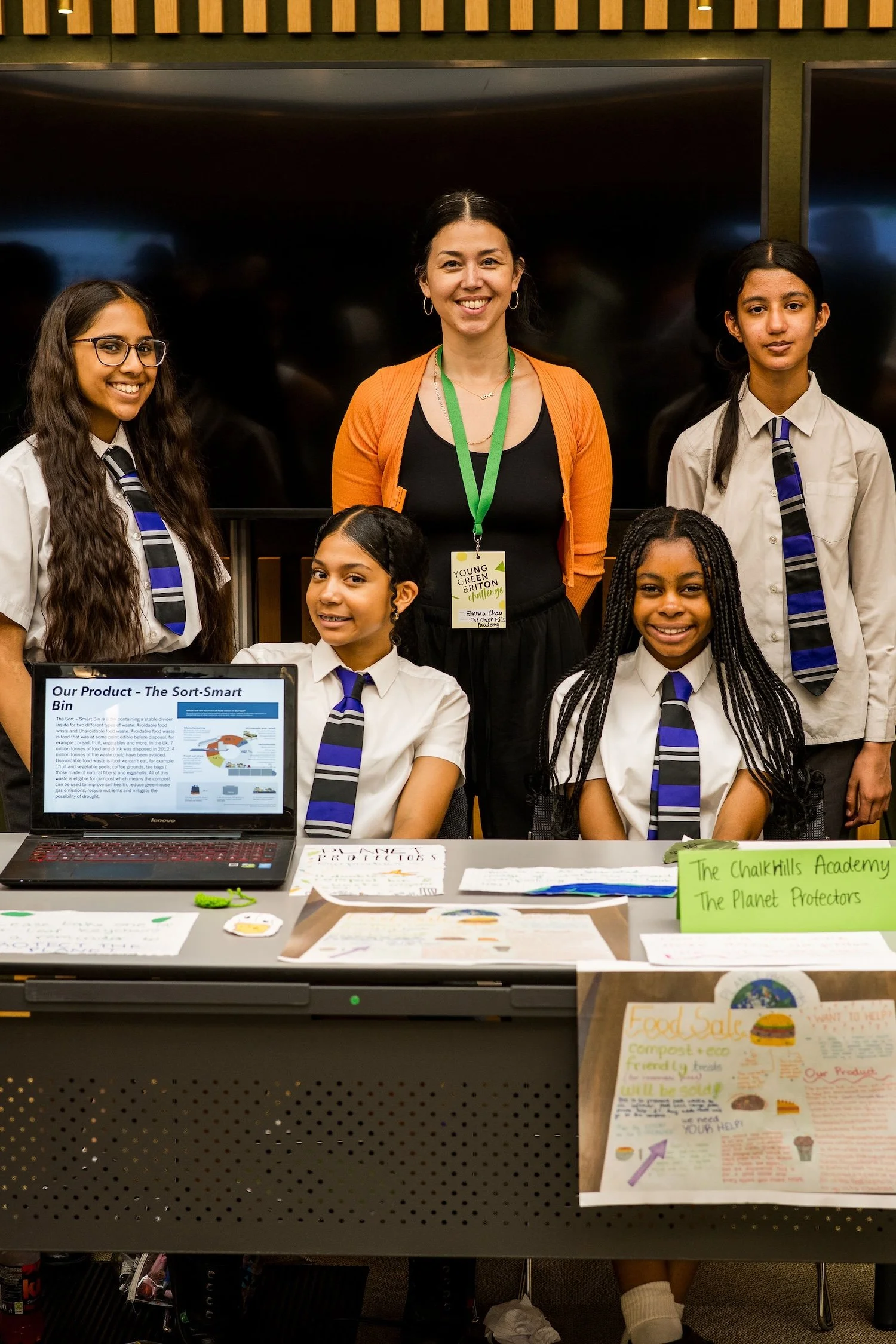 A group of five young girls and one adult woman standing behind a table at a school fair or science exhibition. The women and students are smiling, wearing school uniforms and badges. The table displays various posters, papers, and a laptop. Signage on the table indicates a project called 'The Sort-Smart Bin' and another titled 'The Planet Protectors.' The background includes a large black screen and wooden paneling.