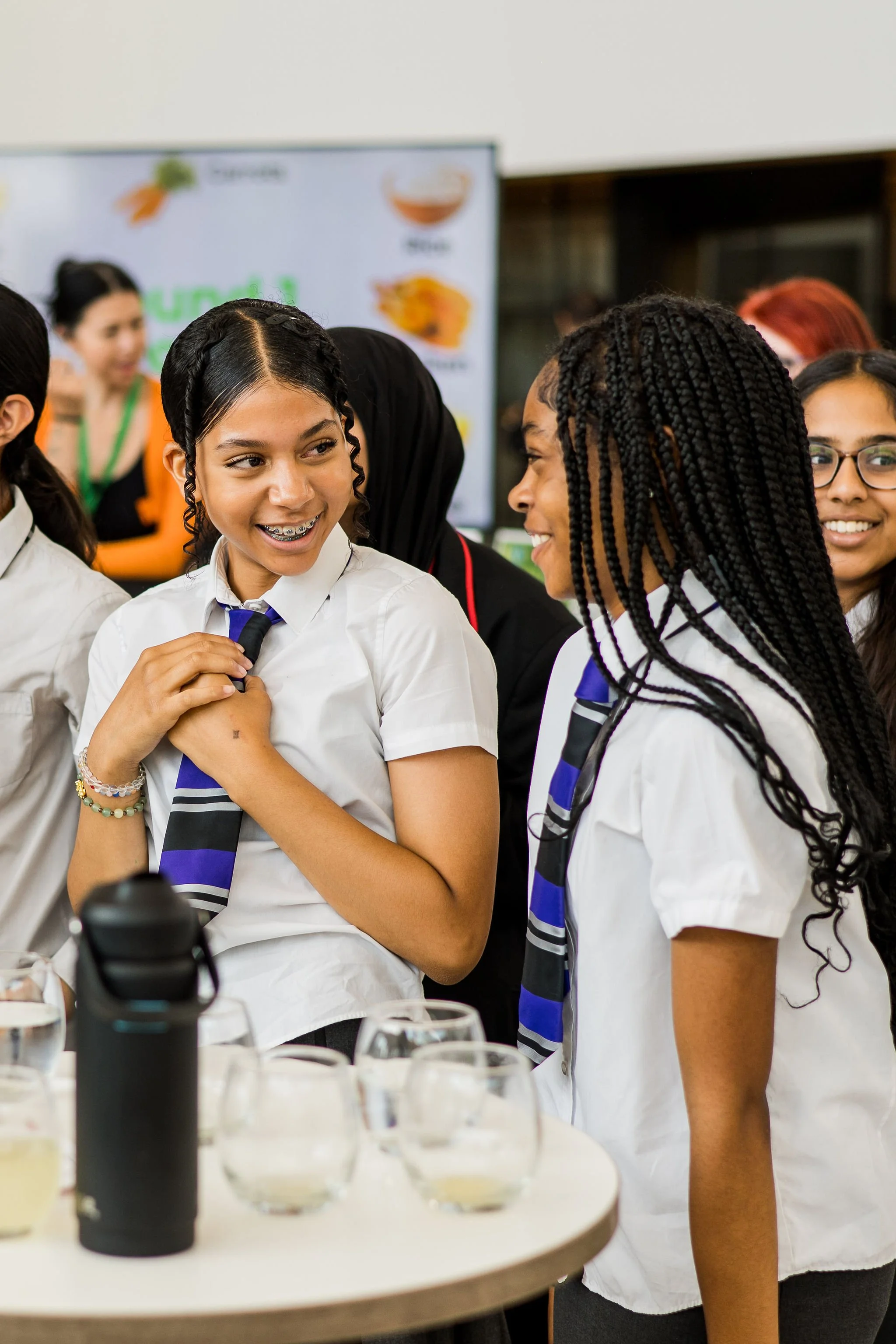 Group of girls in school uniforms smiling and talking at a science event, with test tubes and scientific equipment on the table.