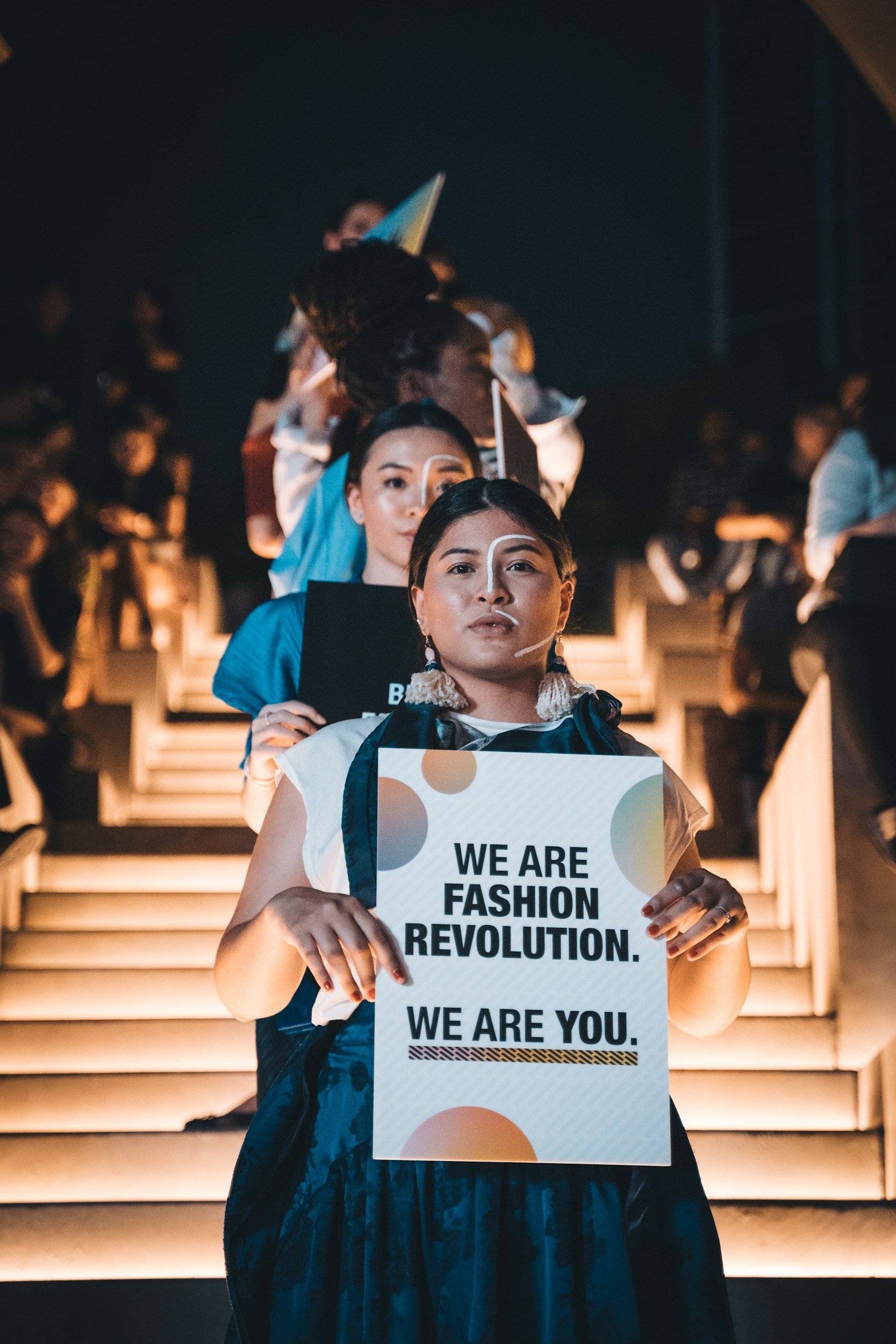 A woman with dark hair holds a sign that reads "We are fashion revolution. We are you." in front of an indoor staircase at a fashion event, with other people visible in the background.