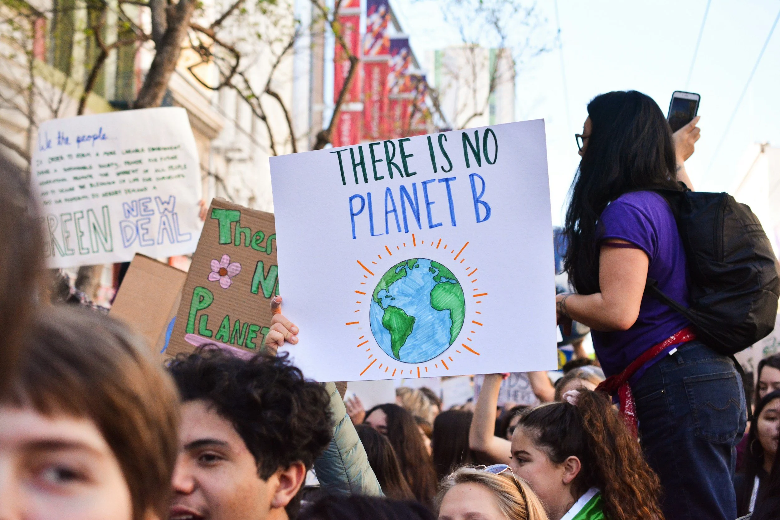 A crowd of people at an outdoor rally or protest, holding signs including one that says 'There is no Planet B' with a drawing of Earth. Another sign visible in the background mentions a green deal and protecting the planet.