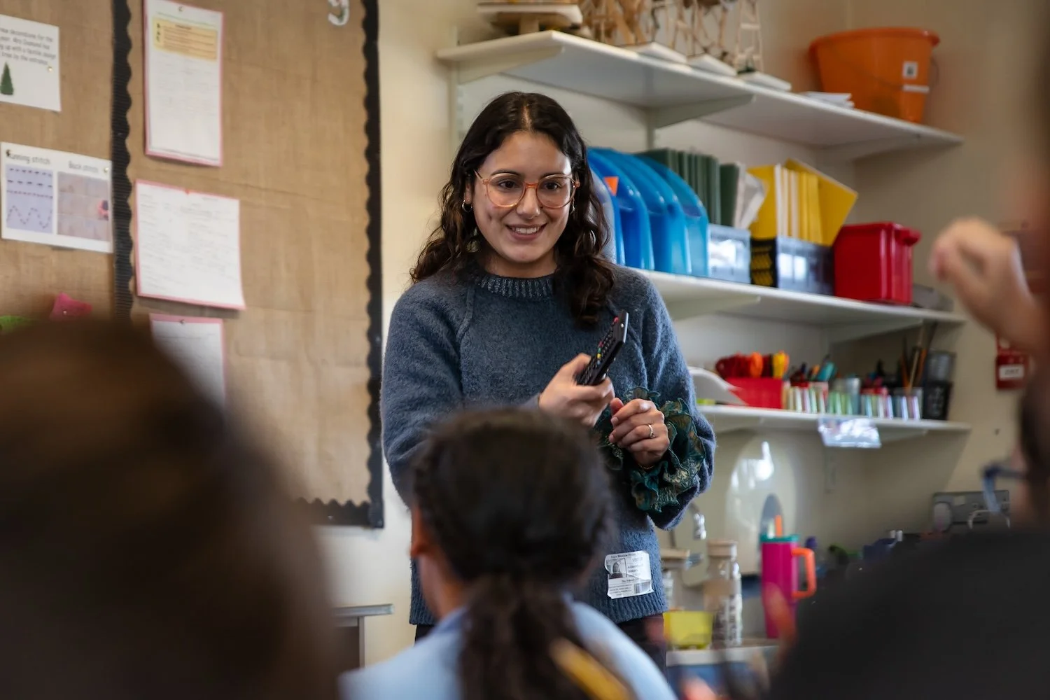 A smiling teacher with glasses holding a remote control, standing in a classroom with students facing her during a lesson.