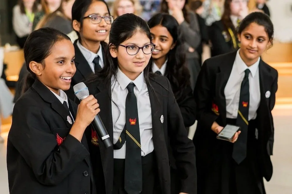 Group of young girls in school uniforms, one speaking into a microphone, listening attentively in a school setting, with other students in the background.