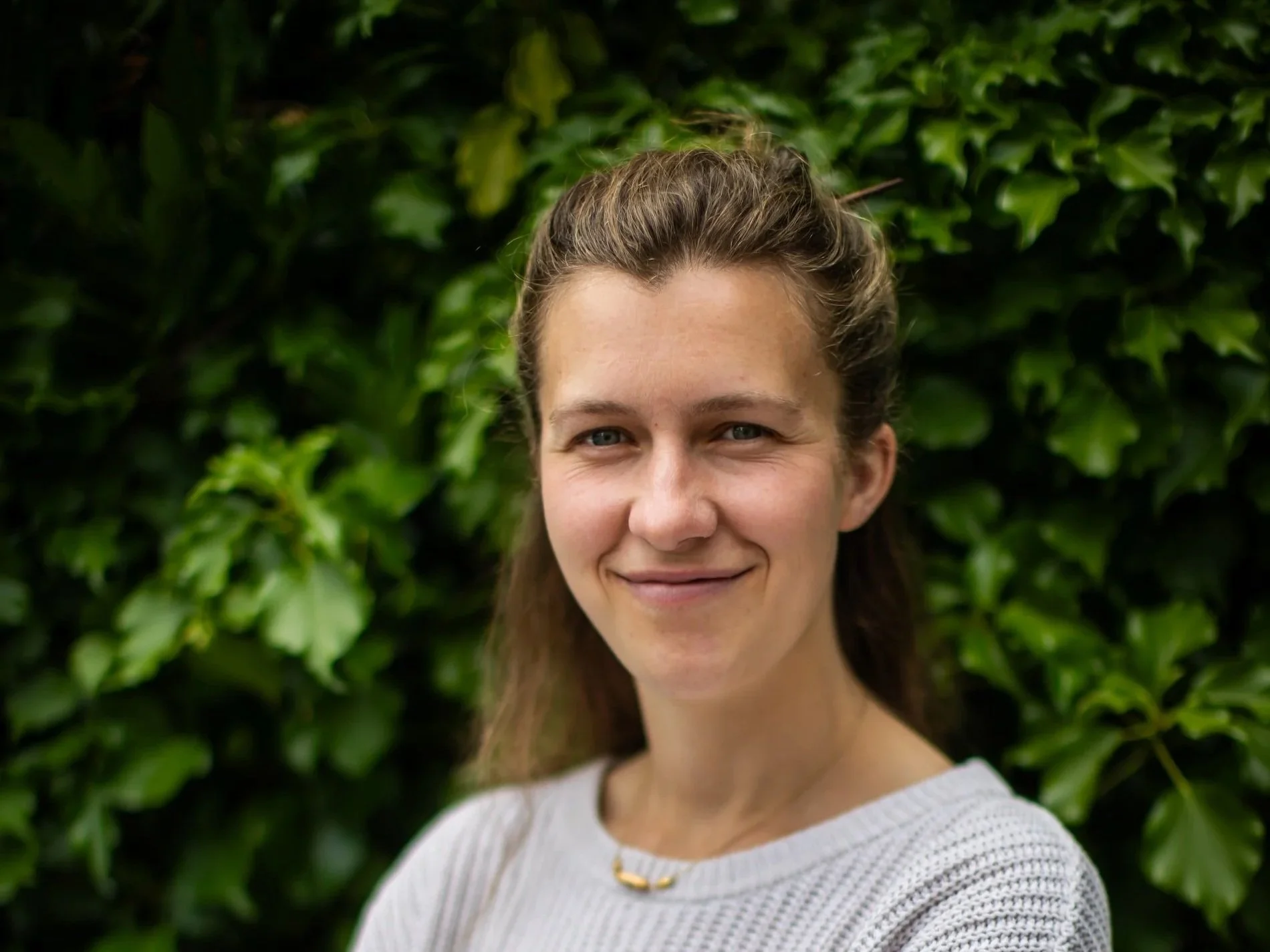 A woman with brown hair and blue eyes smiling outdoors in front of green leafy bushes, wearing a white knitted top and a small gold necklace.
