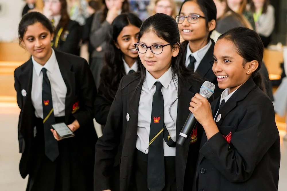 Group of schoolgirls in black uniforms and ties, one holding a microphone, smiling during a school event.
