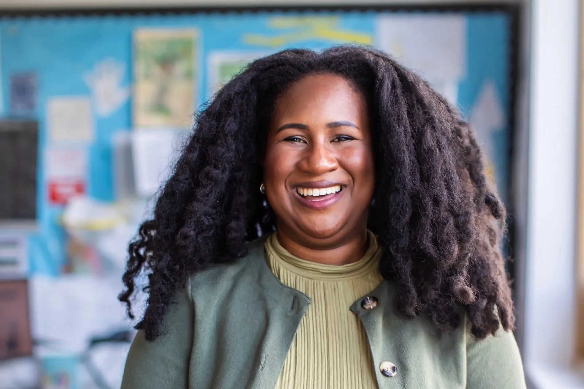 Smiling woman with curly black hair and dark skin standing indoors with a blue bulletin board in the background.